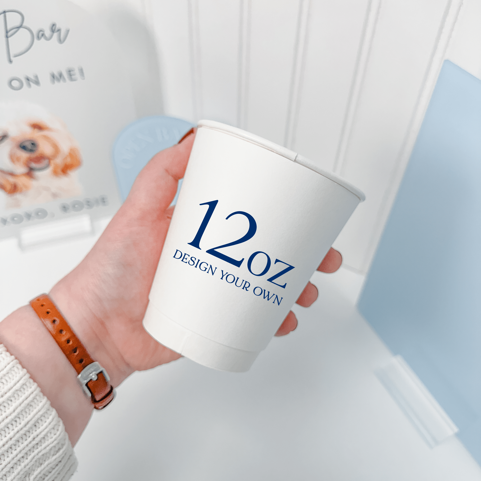 Hand holding a 12 oz white paper cup with a custom navy design, shown for scale