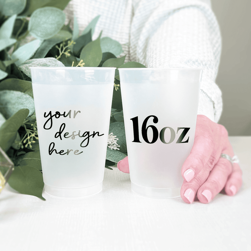 Two frosted 16oz plastic cups with custom text, one reading "your design here" and the other showing "16oz", displayed with greenery and pink decorative pom-poms.
