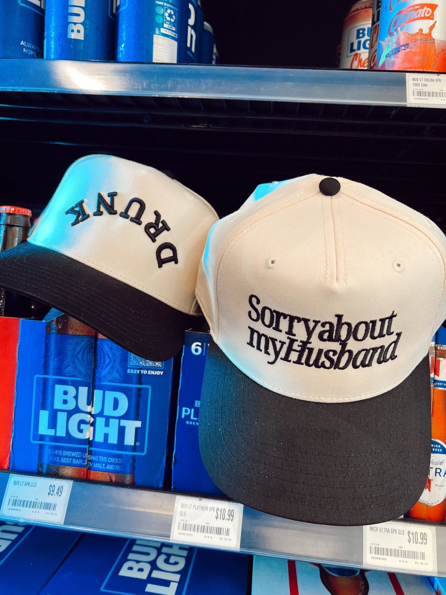 Vintage trucker hat with "Sorry about my Husband" text, cream and black colorway, displayed on store shelf with beverages.