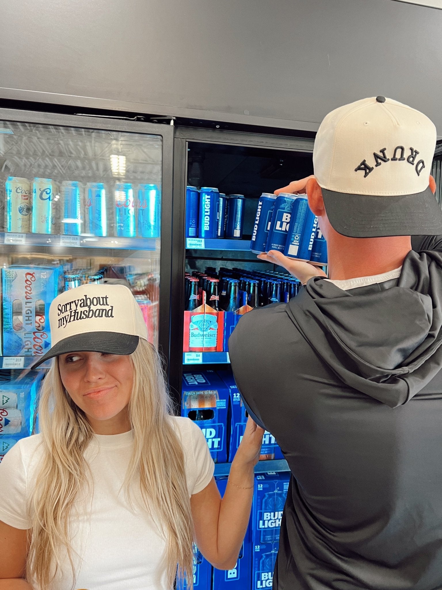 Two people wearing vintage-style trucker hats with curved bills and mesh panels in front of a cooler display, one hat reads "Sorry about my Husband" and the other reads "Sorry about my Wife"