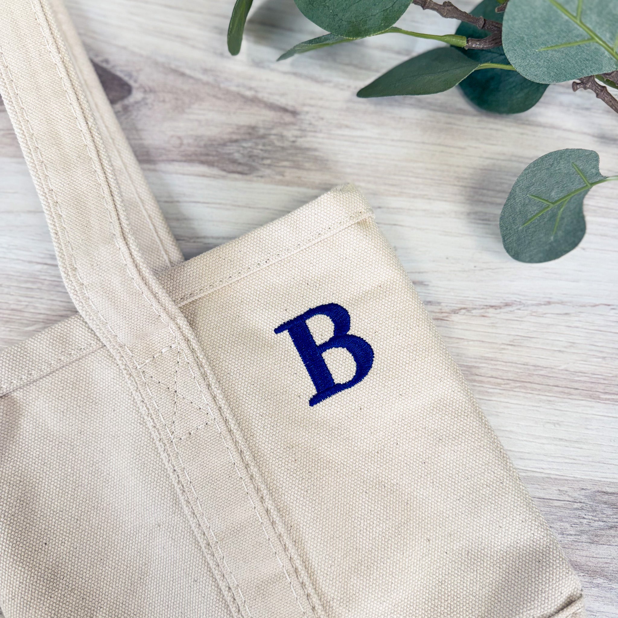 Angled flat lay of monogrammed canvas wine bag showing side panel with embroidered letter and reinforced stitching