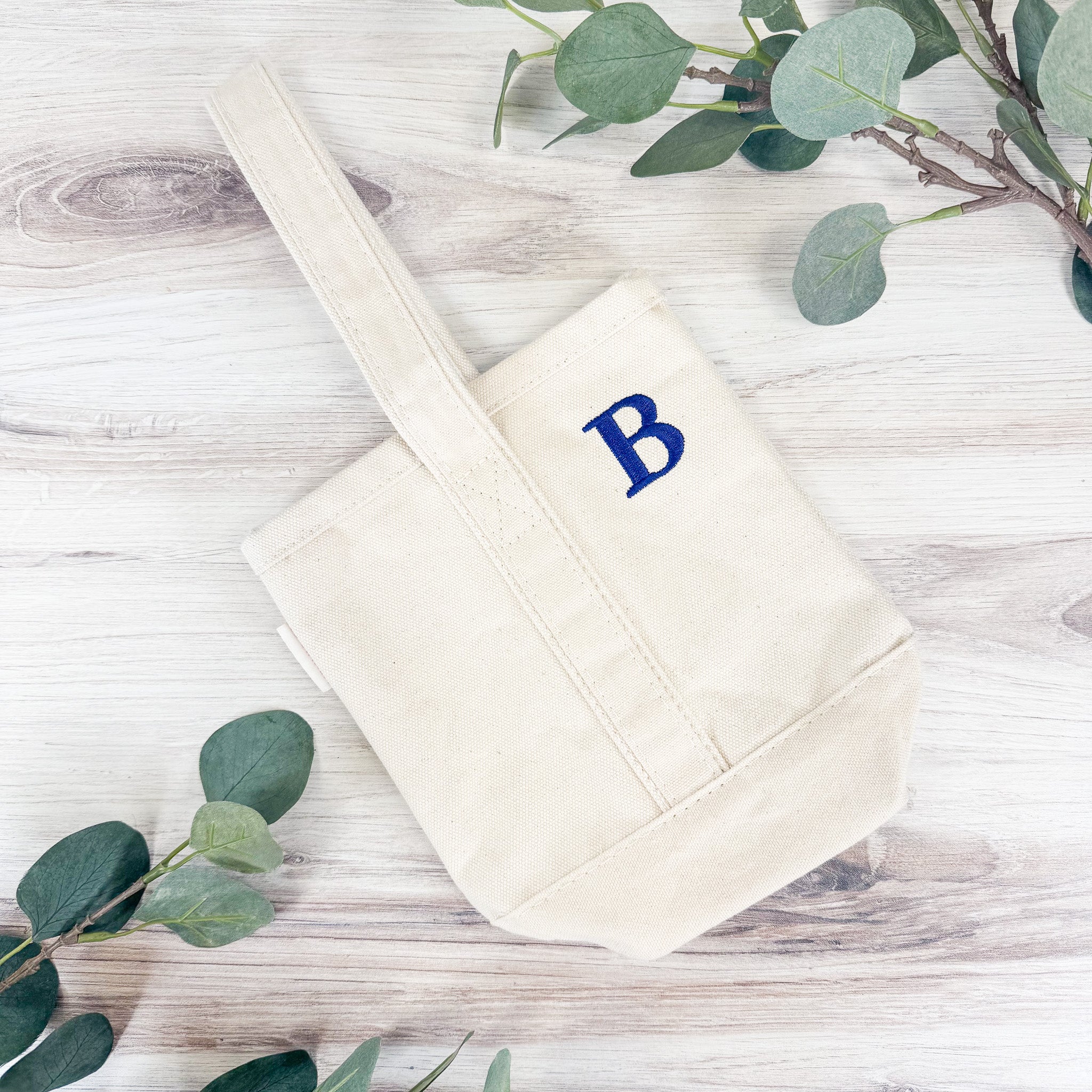 Flat lay of personalized canvas wine tote bag from above, showing embroidered initial and handle, eucalyptus greenery accents