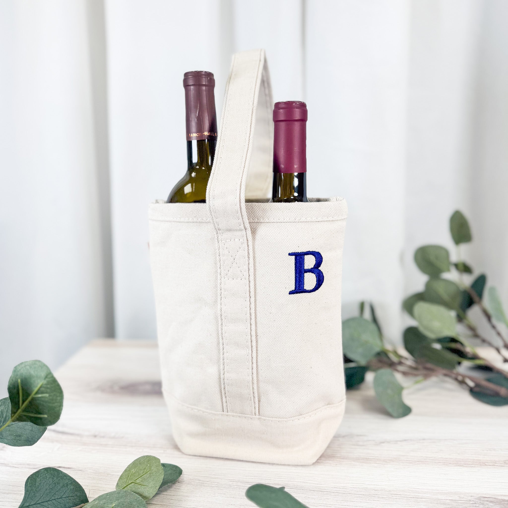 Monogrammed canvas wine carrier with tall handle holding two wine bottles, embroidered B initial in navy blue, eucalyptus styling