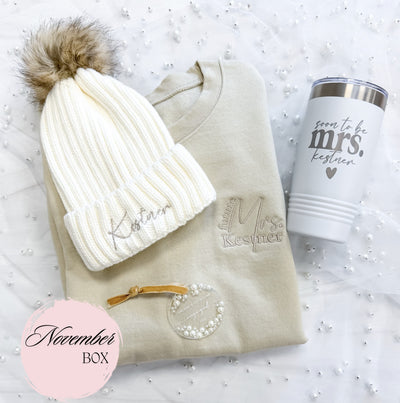 A personalized cozy bridal gift set featuring a cream crewneck sweatshirt, a white ribbed beanie with a faux fur pom-pom, a white insulated tumbler, and a clear acrylic ornament with pearl embellishments, presented for an engagement celebration.