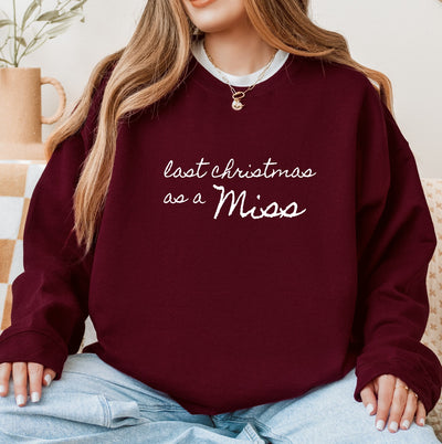 A cozy maroon crewneck sweatshirt made of soft fabric, featuring custom white script text, suitable for a bride-to-be during the Christmas season.