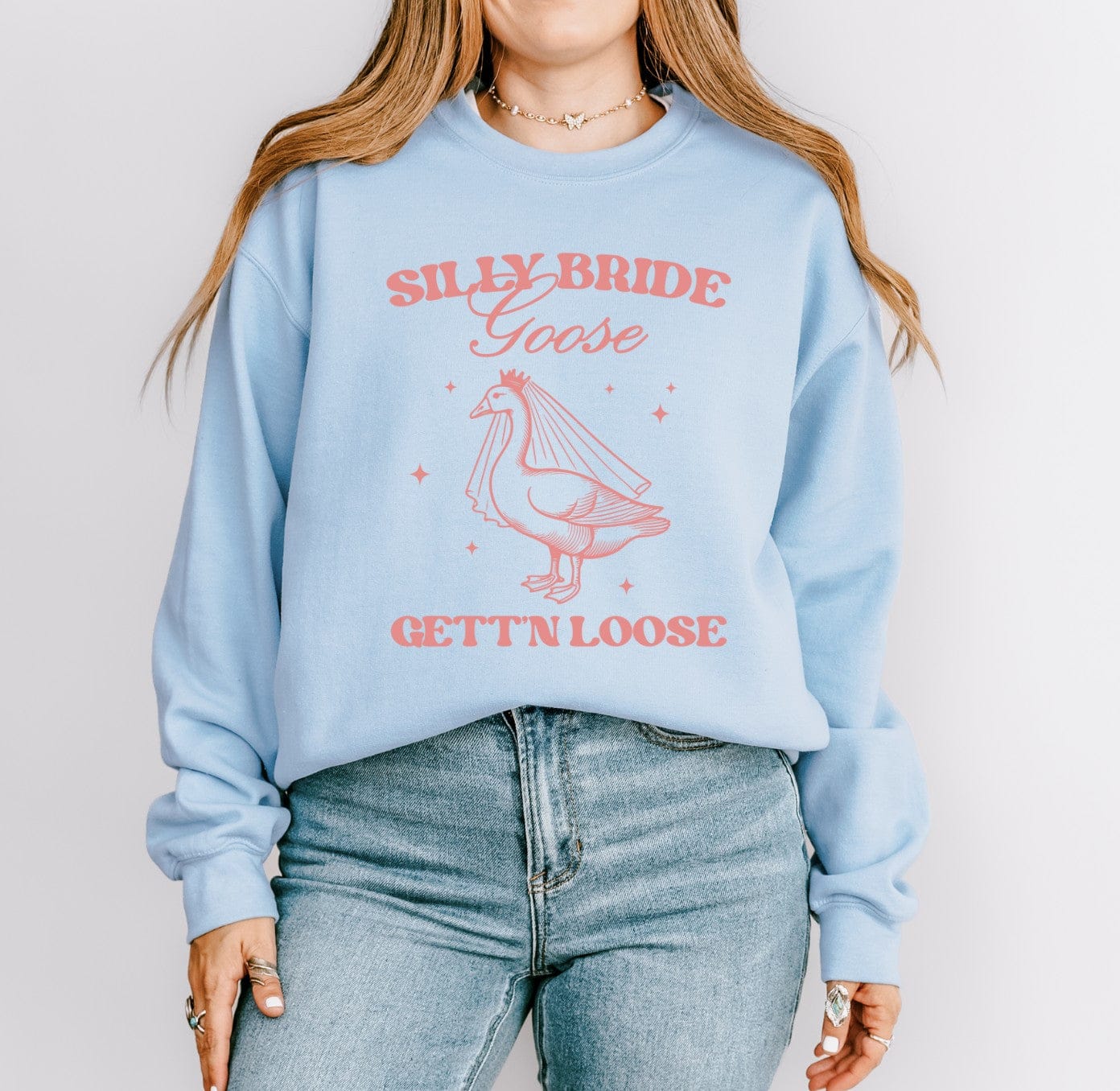 A light blue crewneck sweatshirt featuring a coral pink graphic of a goose wearing a wedding veil and crown, surrounded by sparkling stars, perfect for a bridal event.