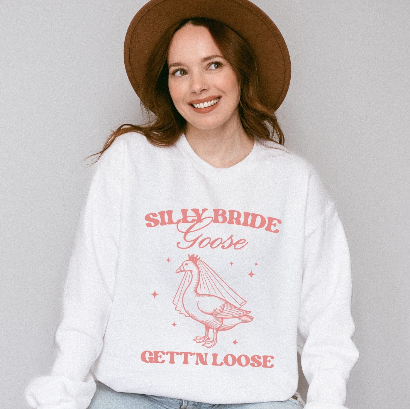 A soft white crewneck sweatshirt featuring a fun pink graphic of a goose wearing a bridal veil and crown, designed for a wedding celebration or bridal shower.