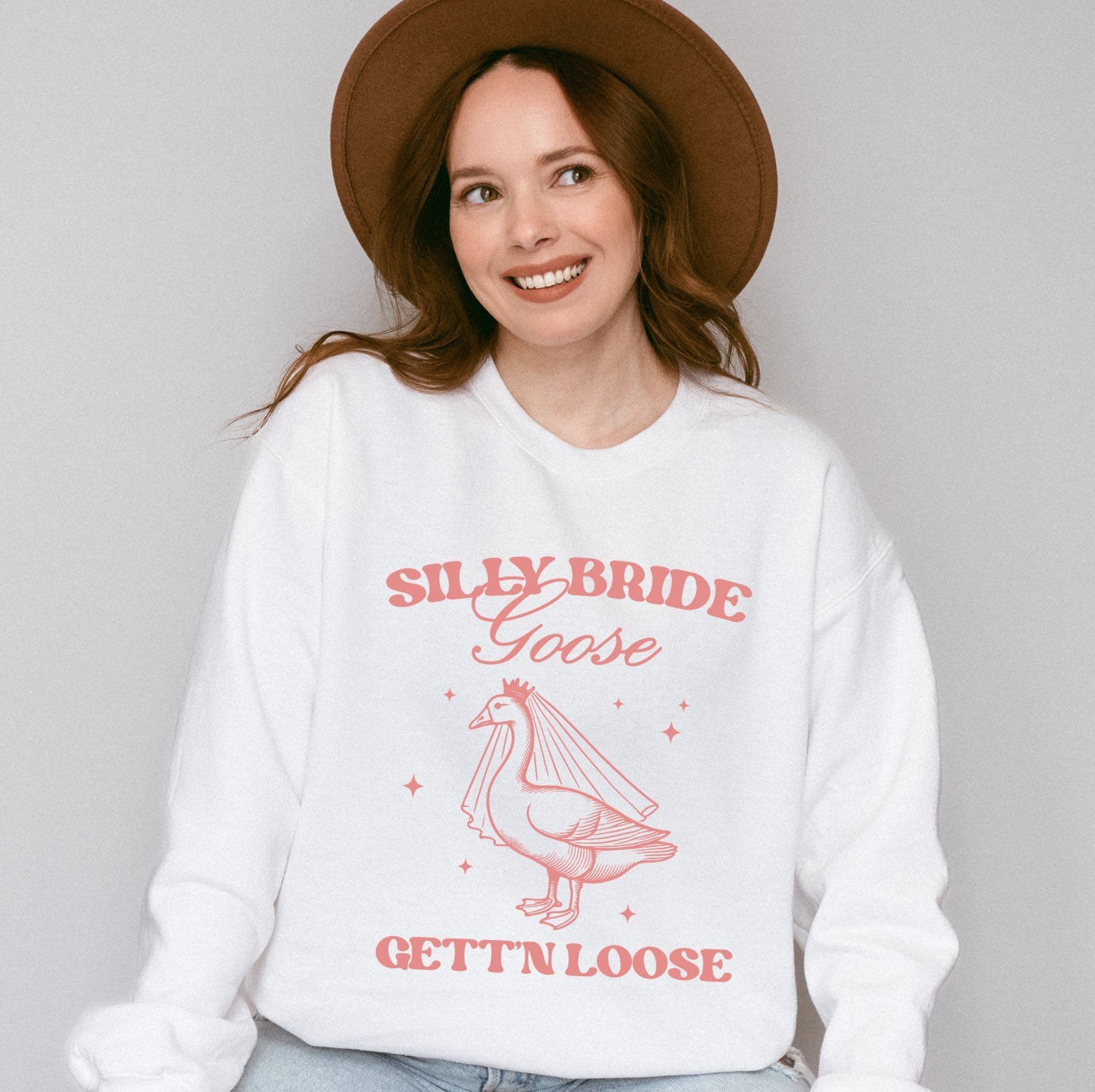 A soft white crewneck sweatshirt featuring a fun pink graphic of a goose wearing a bridal veil and crown, designed for a wedding celebration or bridal shower.
