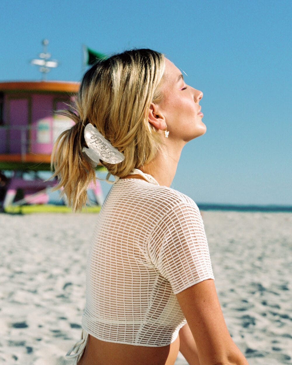 Woman wearing pearlescent claw clip in blonde hair at beach with colorful lifeguard stand in background.