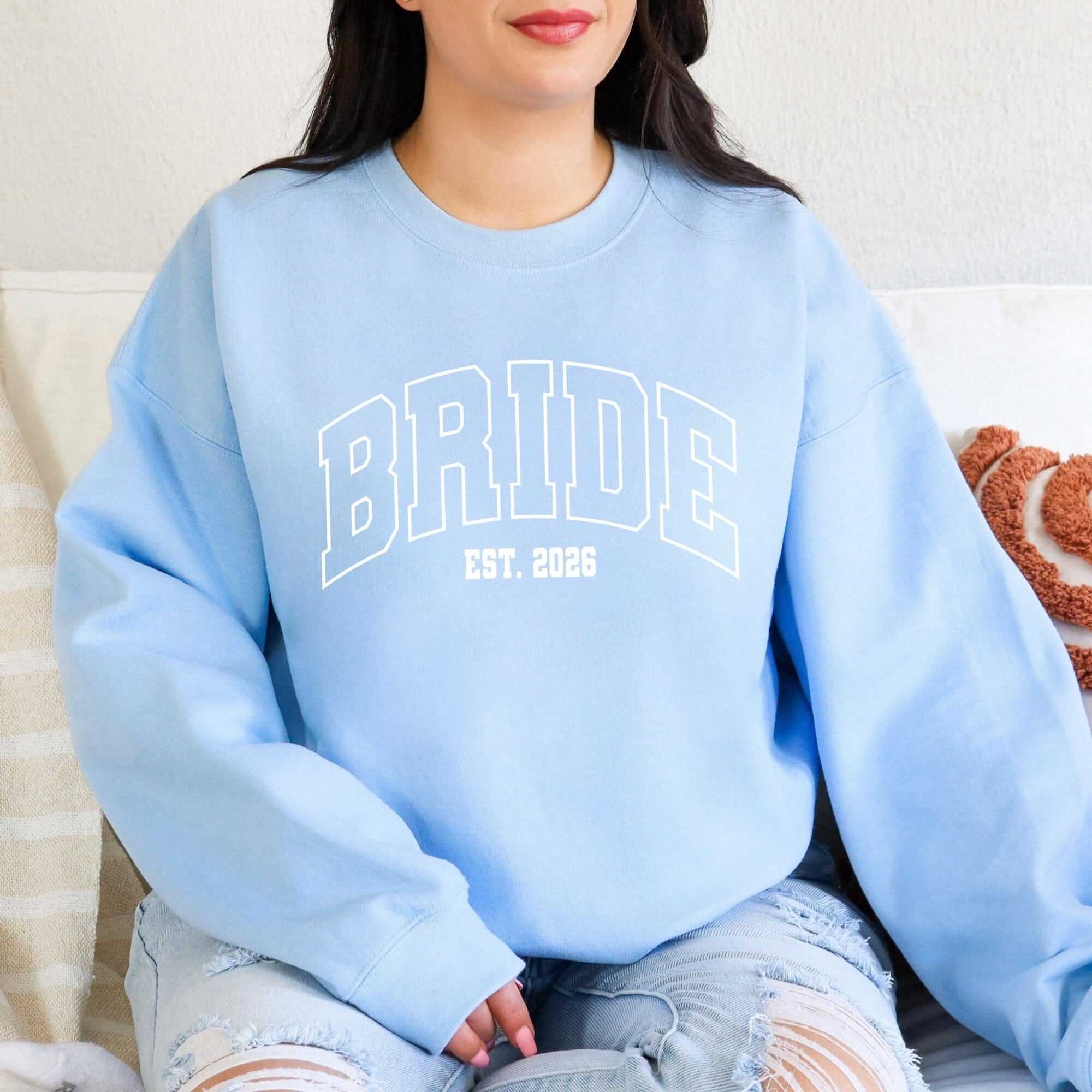 A light blue crewneck sweatshirt made from soft fabric features white bold arched lettering, perfect for a bridal event or celebration.