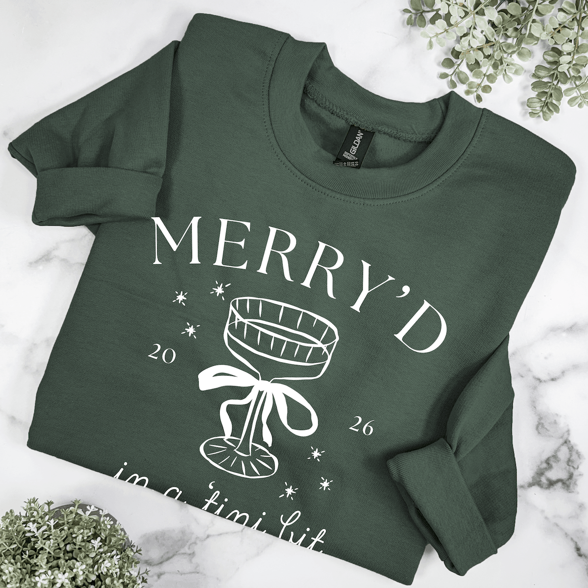 Folded forest green Christmas bride crewneck sweatshirt with Merry'd in a Tini Bit design, white champagne glass graphic, and 2026 date.