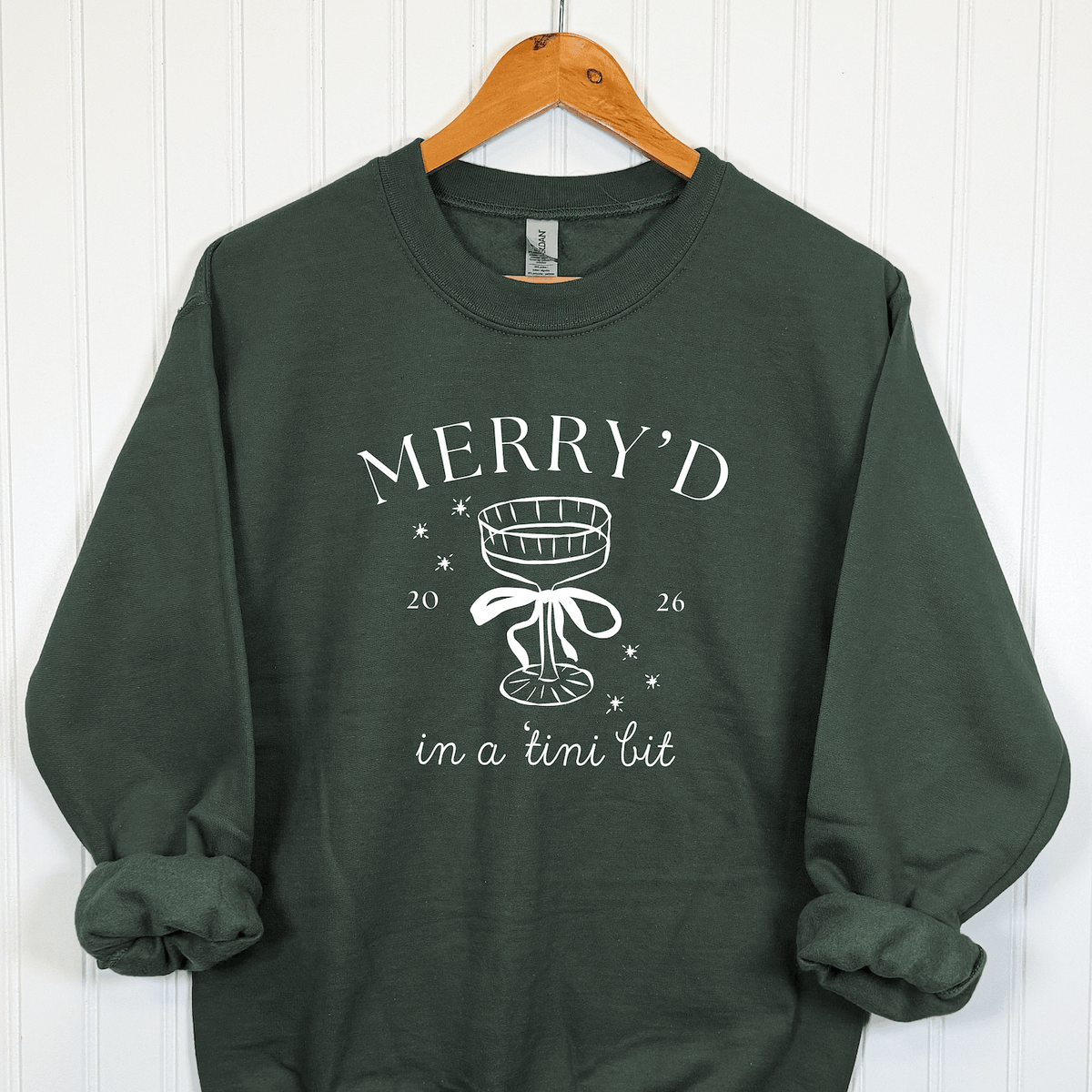 Forest green Merry'd in a Tini Bit Christmas bride sweatshirt featuring white champagne coupe illustration, bow, snowflakes, and festive bridal lettering.