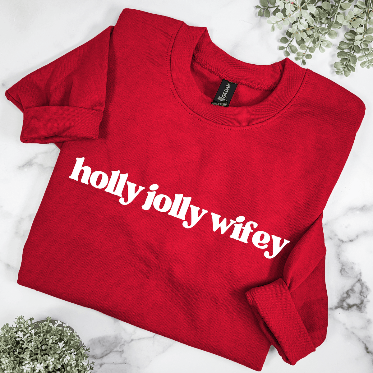 Red Holly Jolly Wifey Christmas sweatshirt featuring bold white retro lettering, folded on a marble background with greenery for a festive holiday look.