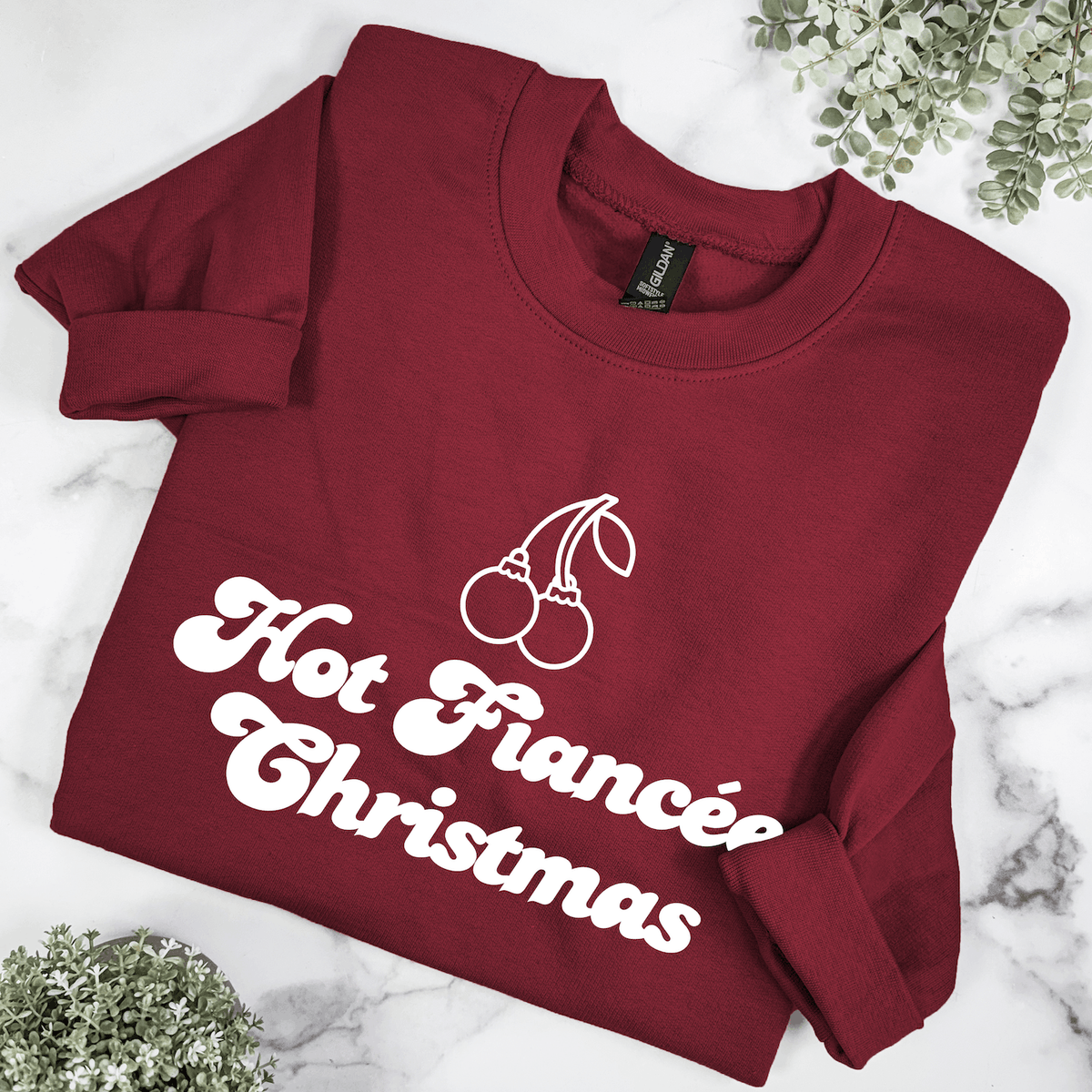 Burgundy Hot Fiancée Christmas sweatshirt featuring white retro lettering and cherry ornament illustration, folded on a marble background with greenery.