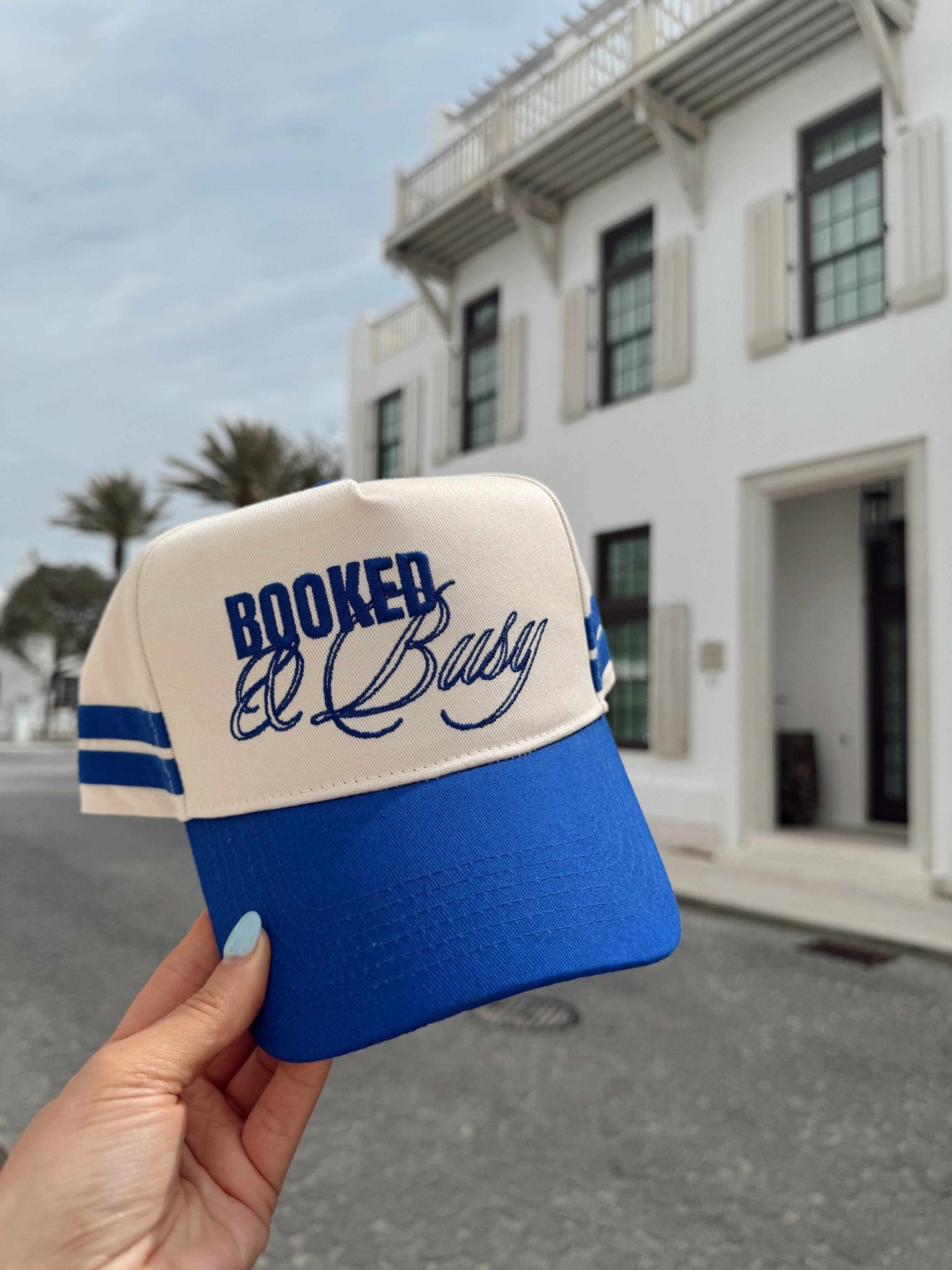 Blue and white vintage trucker hat with "Booked and Busy" text and striped detail, held in hand against white building backdrop.