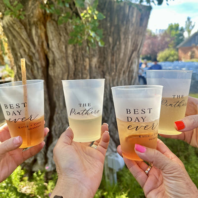 Four frosted clear plastic cups, each containing a beverage, are held outdoors, showcasing personalized designs perfect for a wedding or special event.