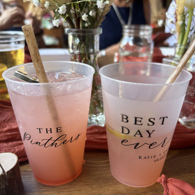 Two translucent plastic cups, filled with pink beverages and ice, each with a straw, are custom printed for a wedding or special event.
