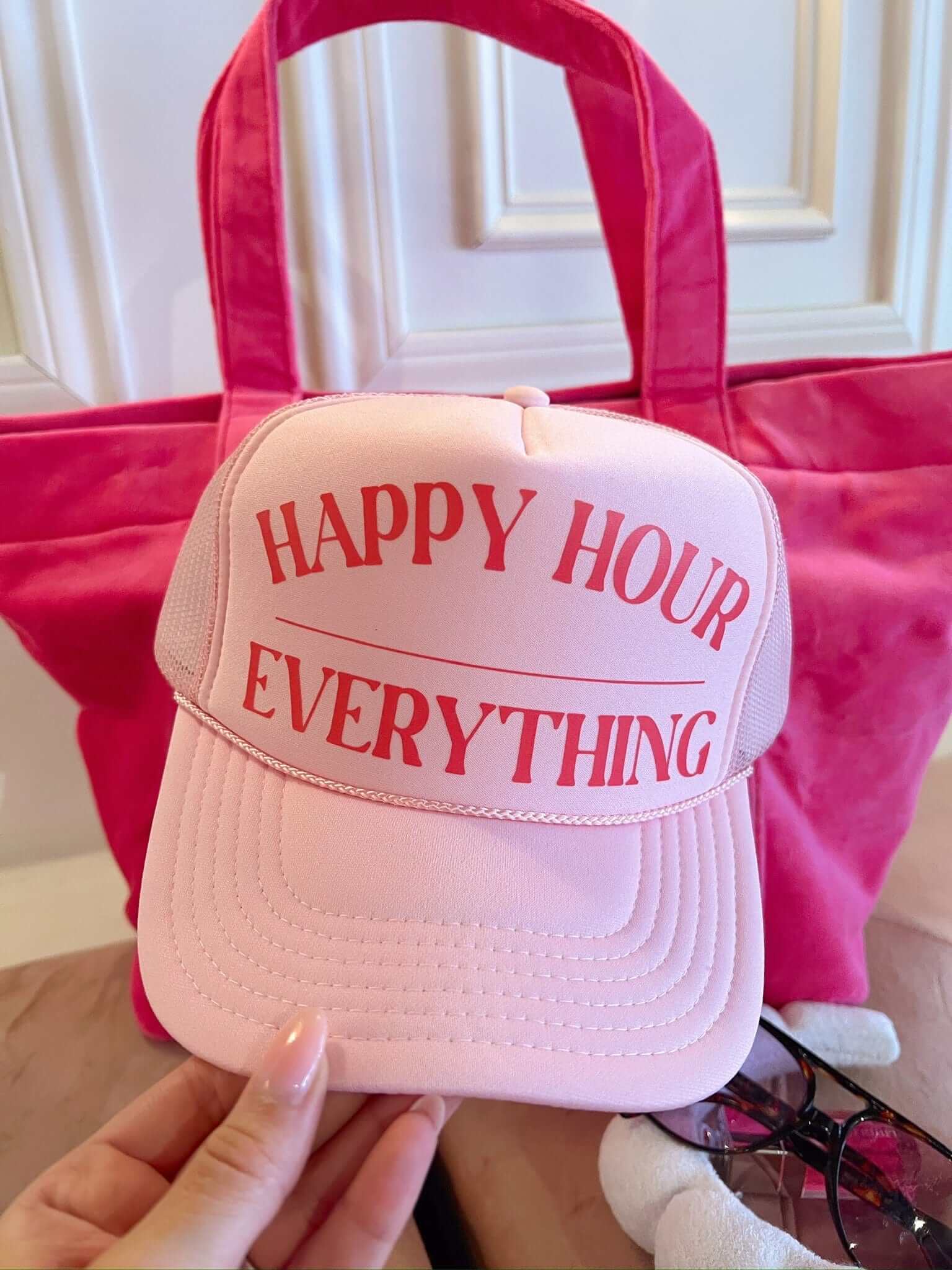 Light pink trucker hat with red "Happy Hour Everything" text, displayed in front of a hot pink tote bag