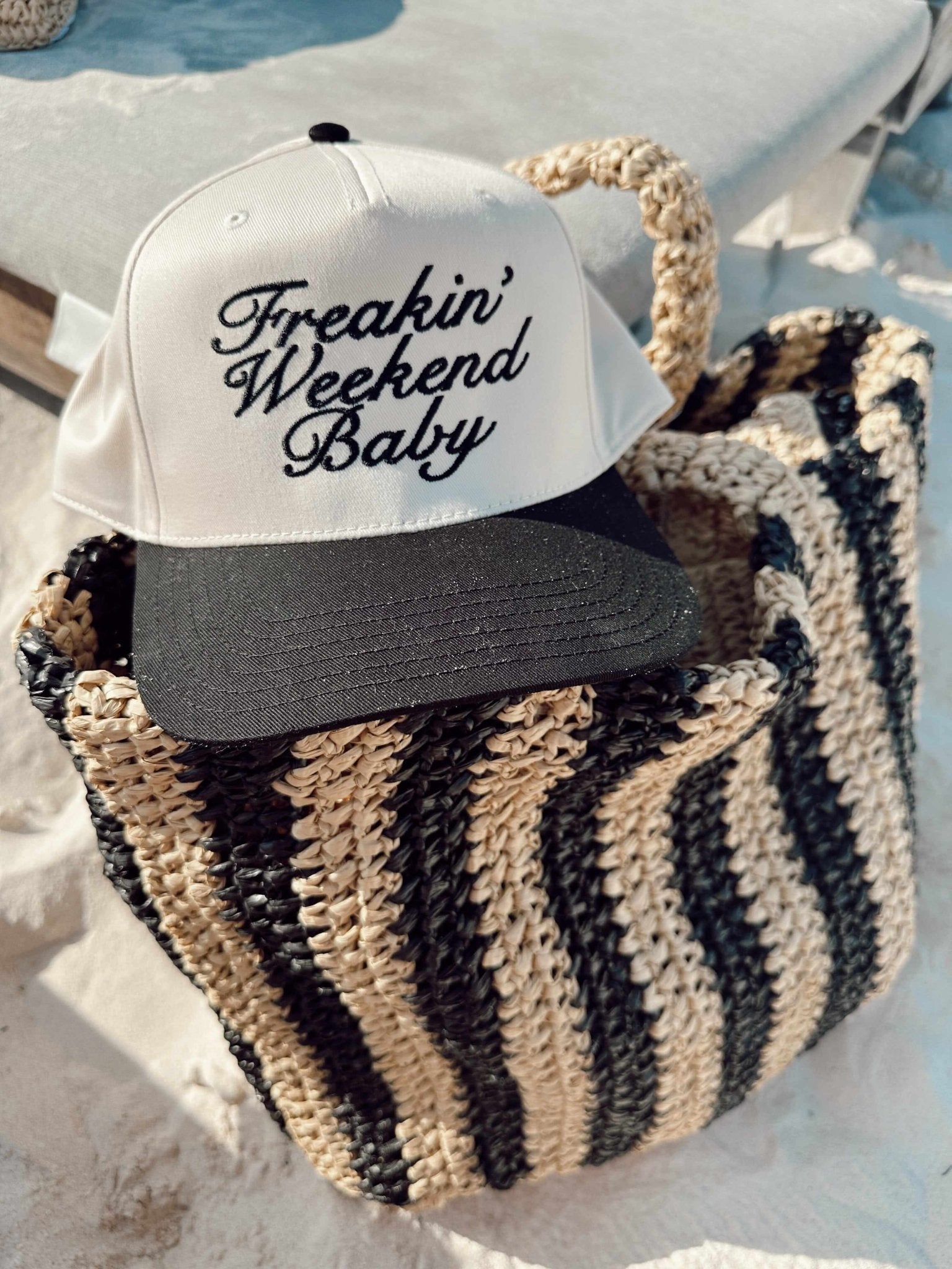 White and black trucker hat with "Freakin' Weekend Baby" script text, resting in a striped woven beach bag.