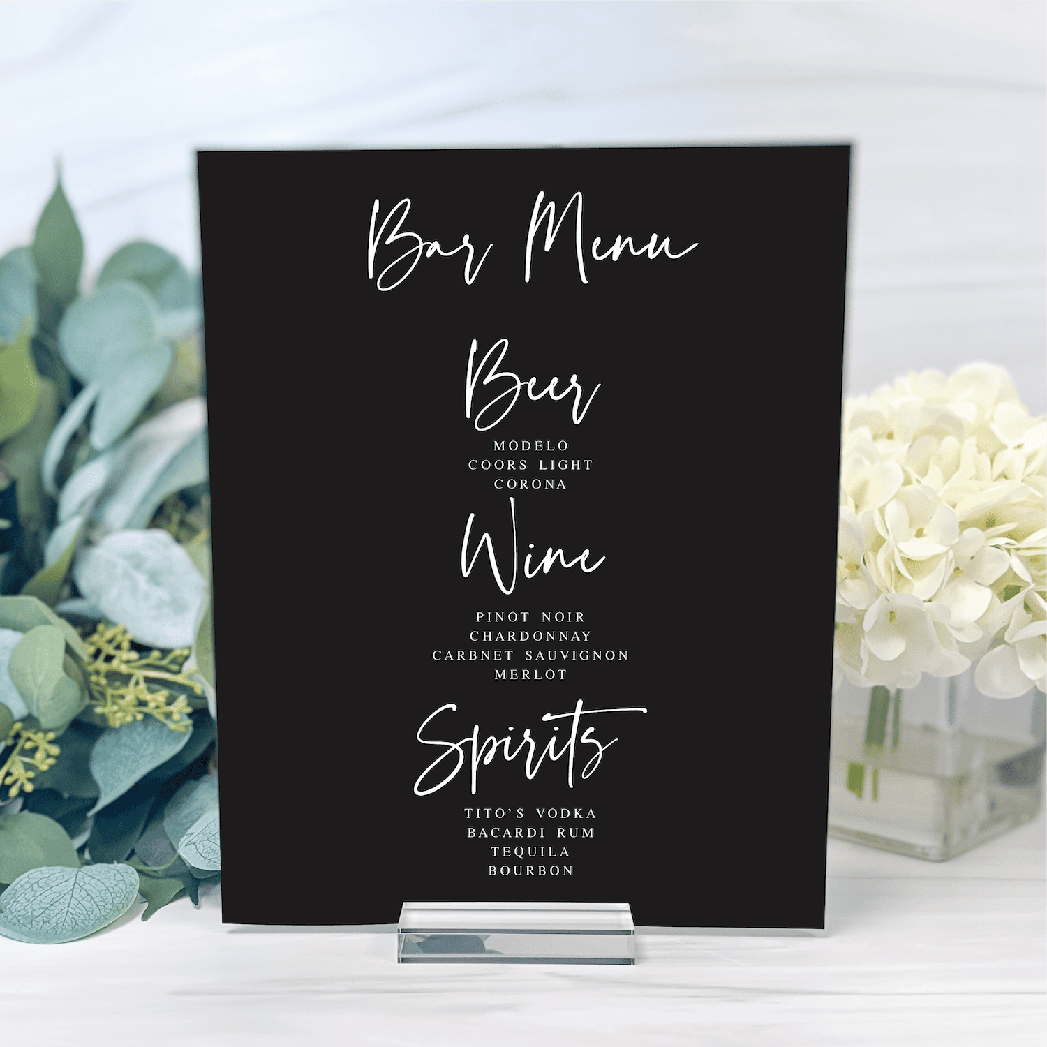 Black bar menu sign with white handwritten-style text listing beer, wine, and spirits options, displayed with easel stand between greenery and white flowers.