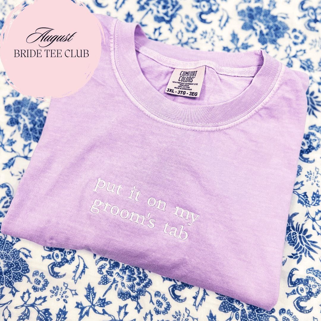 Lavender t-shirt with white embroidered text reading "put it on my grooms tab" displayed on blue floral background