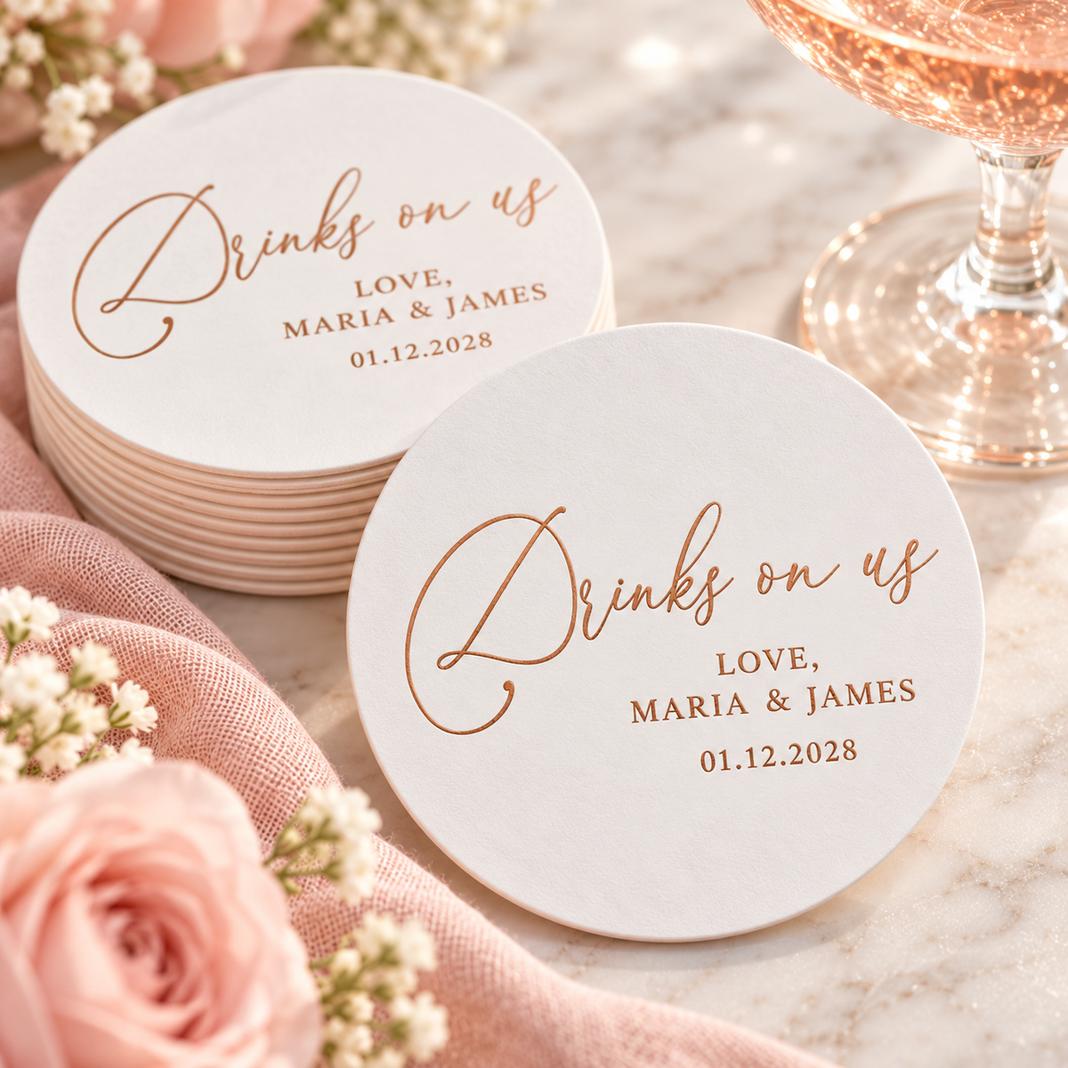 Personalized Drinks on Us wedding coasters with rose gold foil lettering, custom couple names and wedding date, displayed with blush flowers and champagne.
