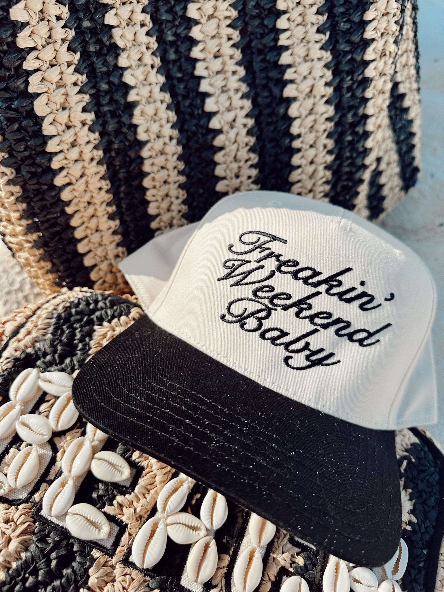 Black and cream vintage trucker hat with "Freakin' Weekend Baby" text, displayed on striped knit fabric with shell embellishments.