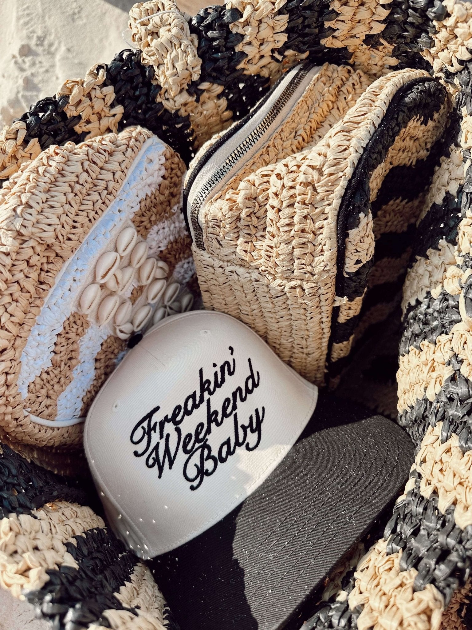 White ceramic mug with "Freakin' Weekend Baby" script text, displayed with straw hats and woven bags on textured fabric.