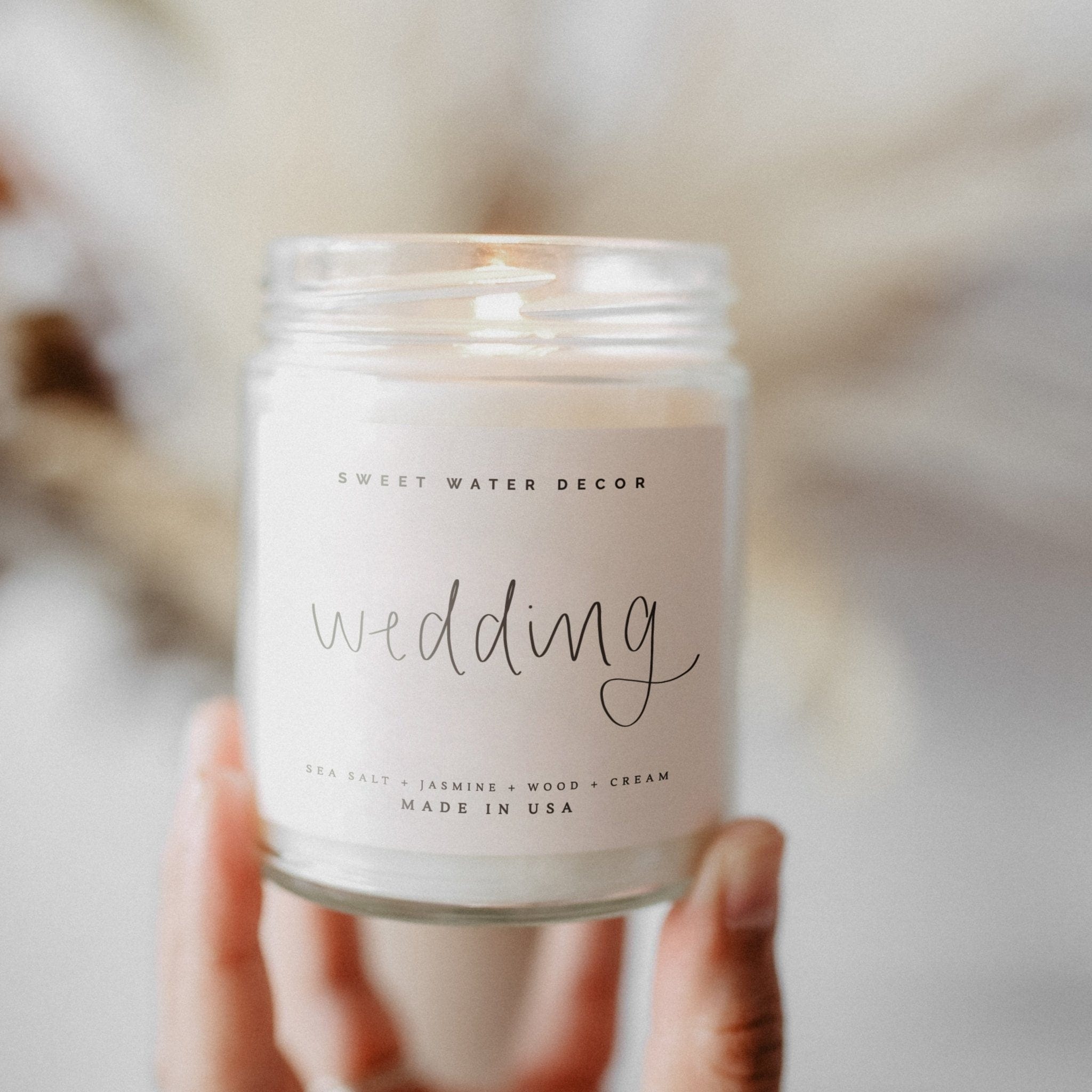 Clear glass jar candle with "wedding" text in script, held in hands against neutral background