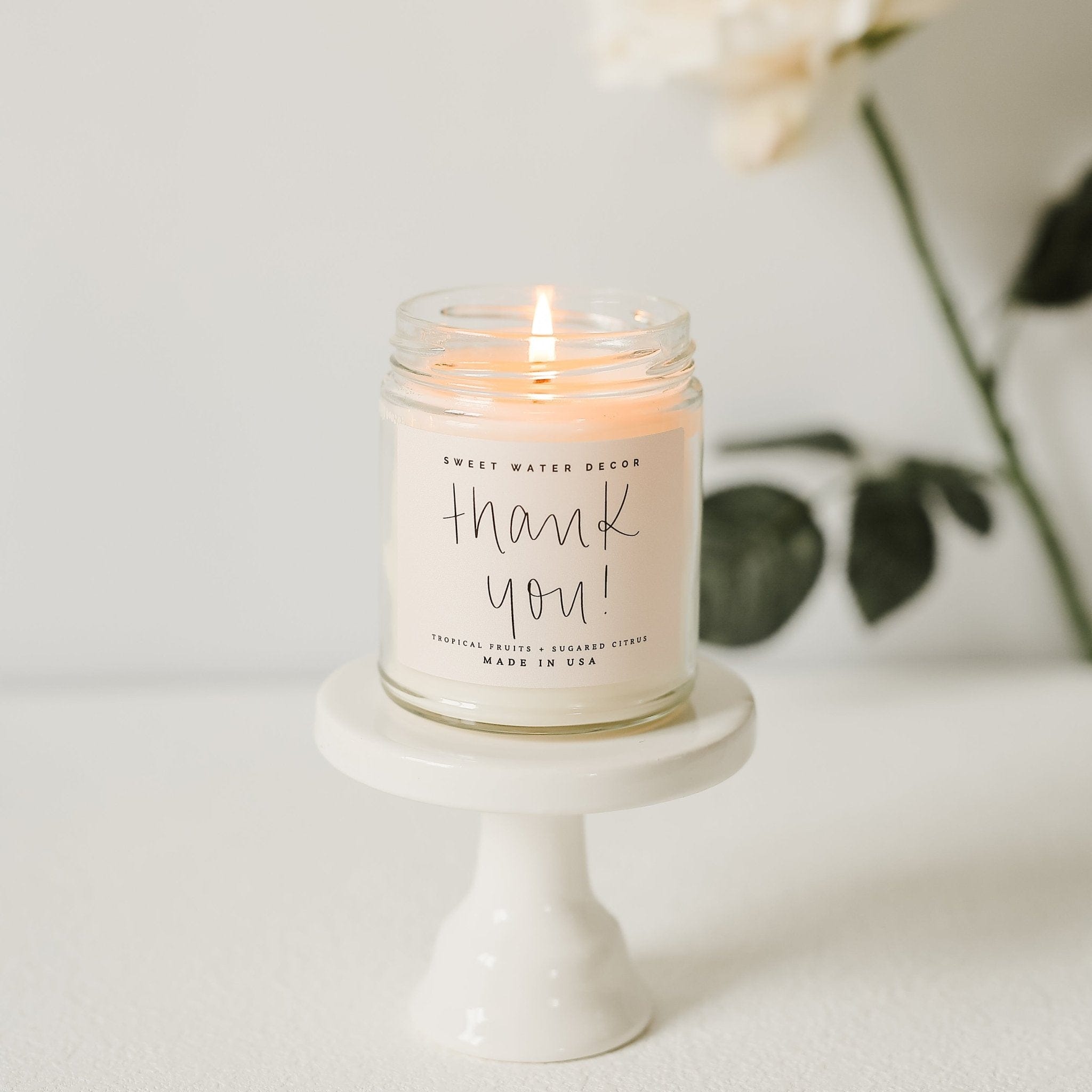 Lit soy candle in clear glass jar with "thank you" handwritten text on cream label, displayed on white pedestal base.