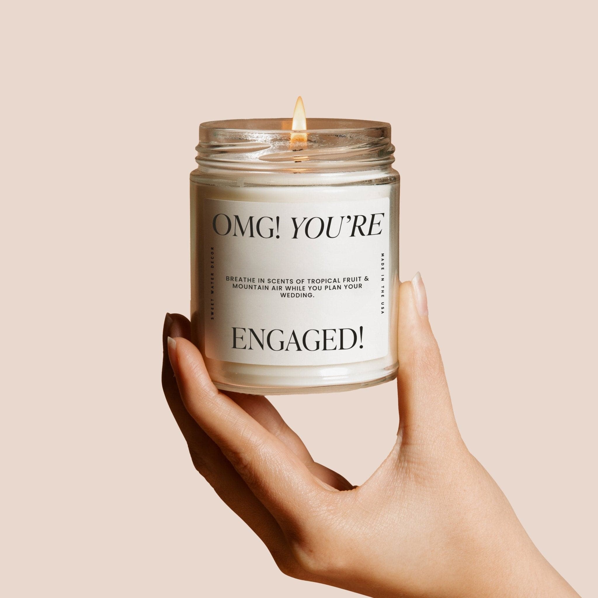 Hand holding lit soy candle in clear glass jar with "OMG! YOU'RE ENGAGED!" text on white label.