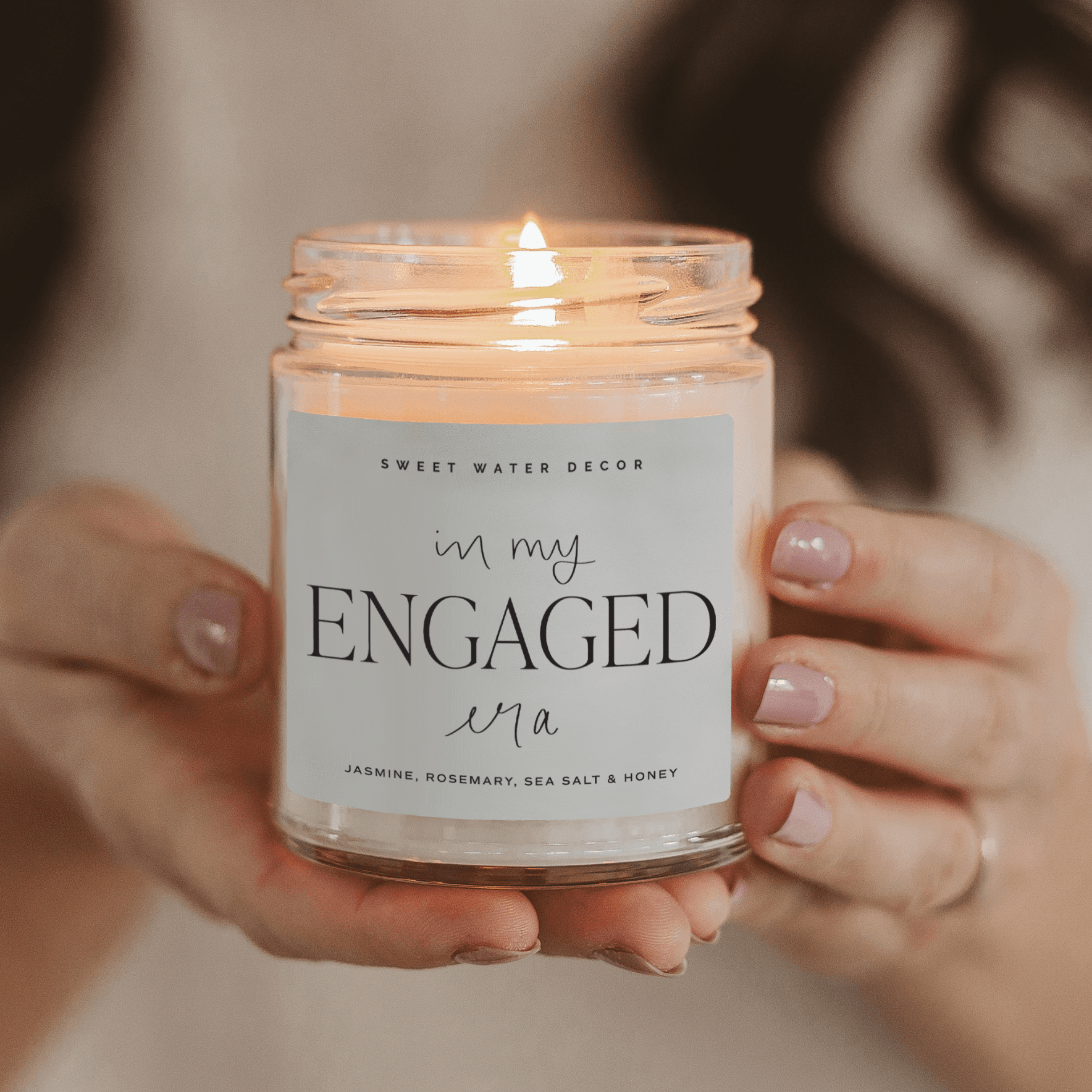 Clear glass jar candle with white label reading "in my ENGAGED era" held in hands with manicured nails
