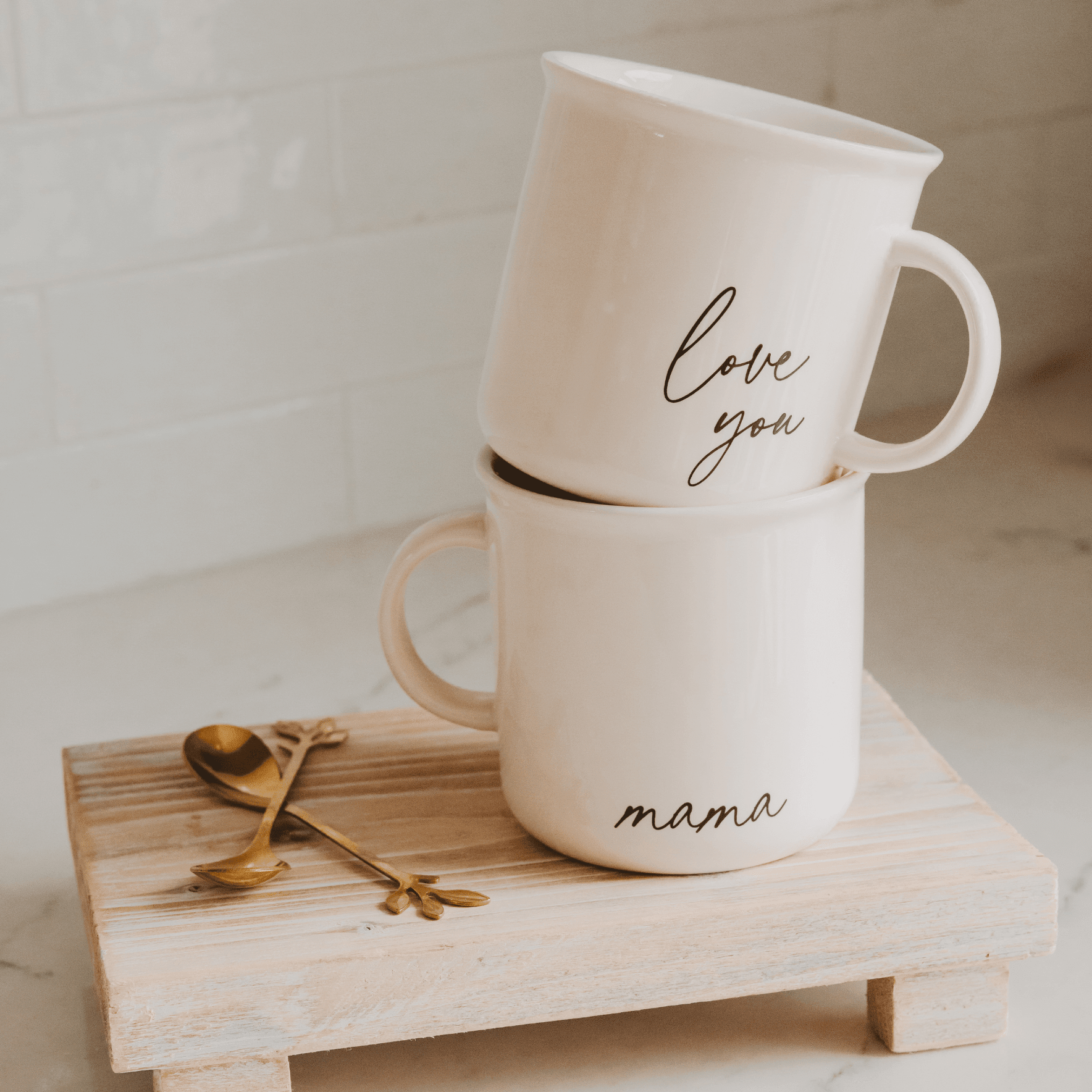 Two stacked white ceramic mugs with "love you" and "mama" text, displayed on a wooden tray with gold decorative leaf accents.