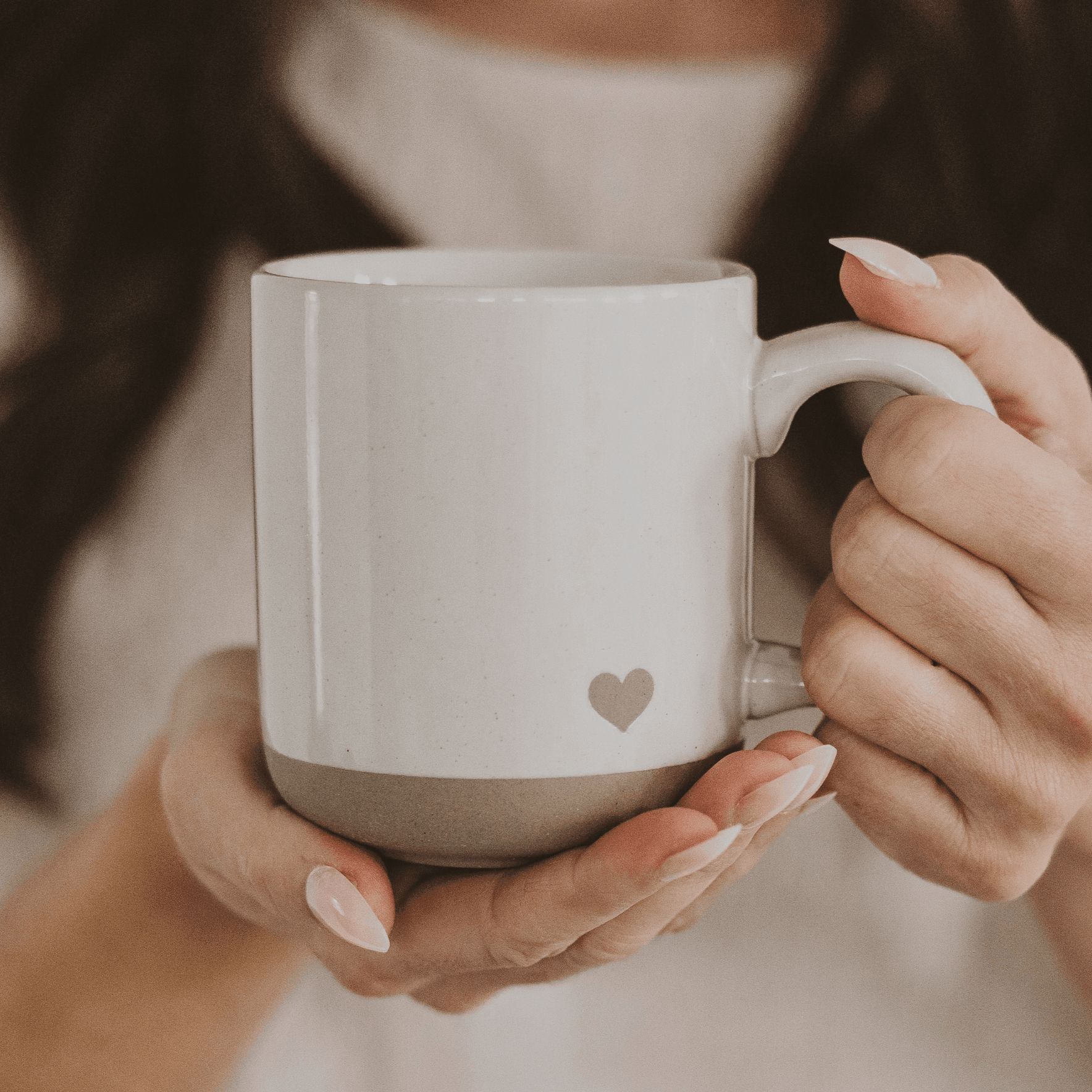 White stoneware mug with small gray heart design, held in hands with natural nails