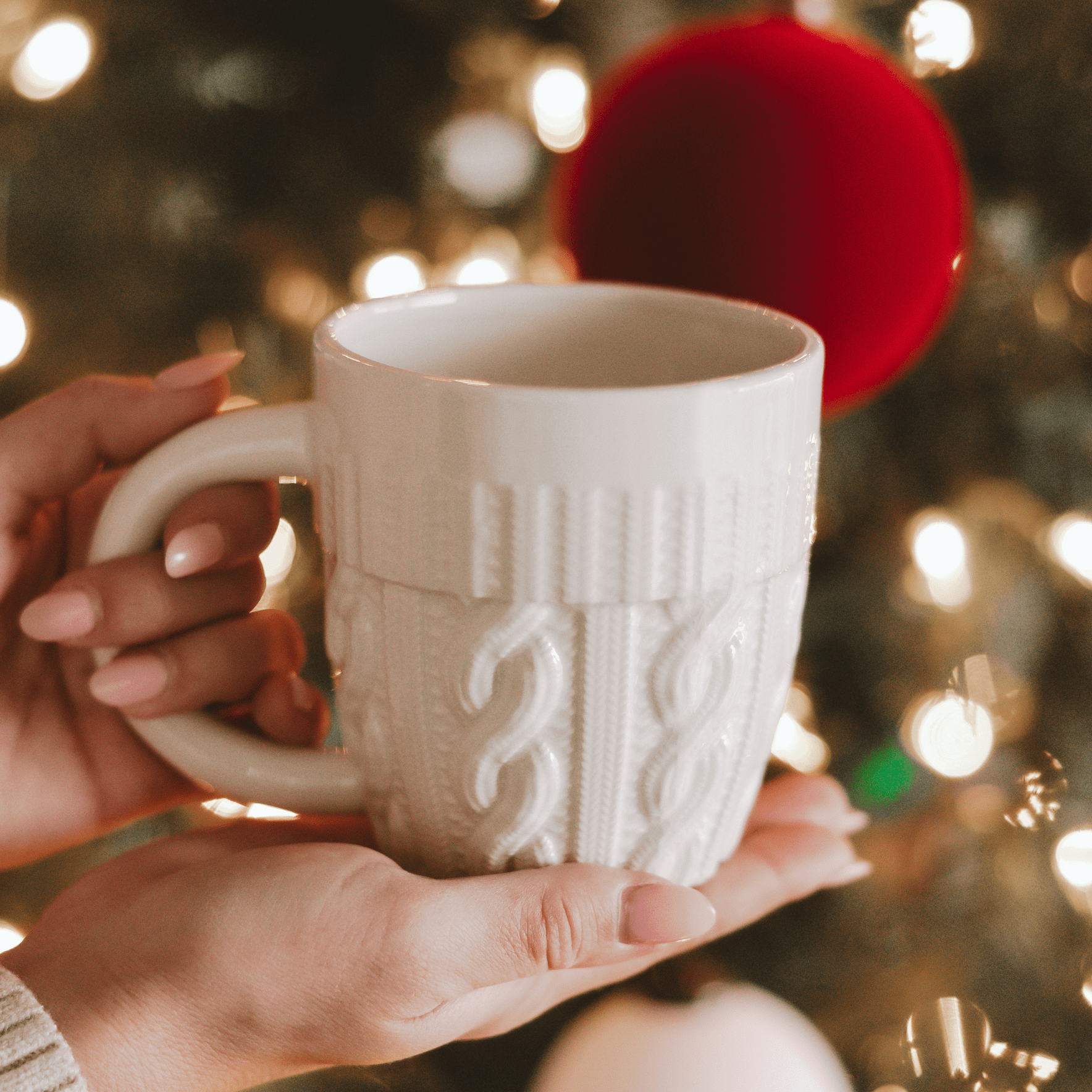 Cream ceramic mug with cozy knit sweater texture design held in hands against holiday lights backdrop