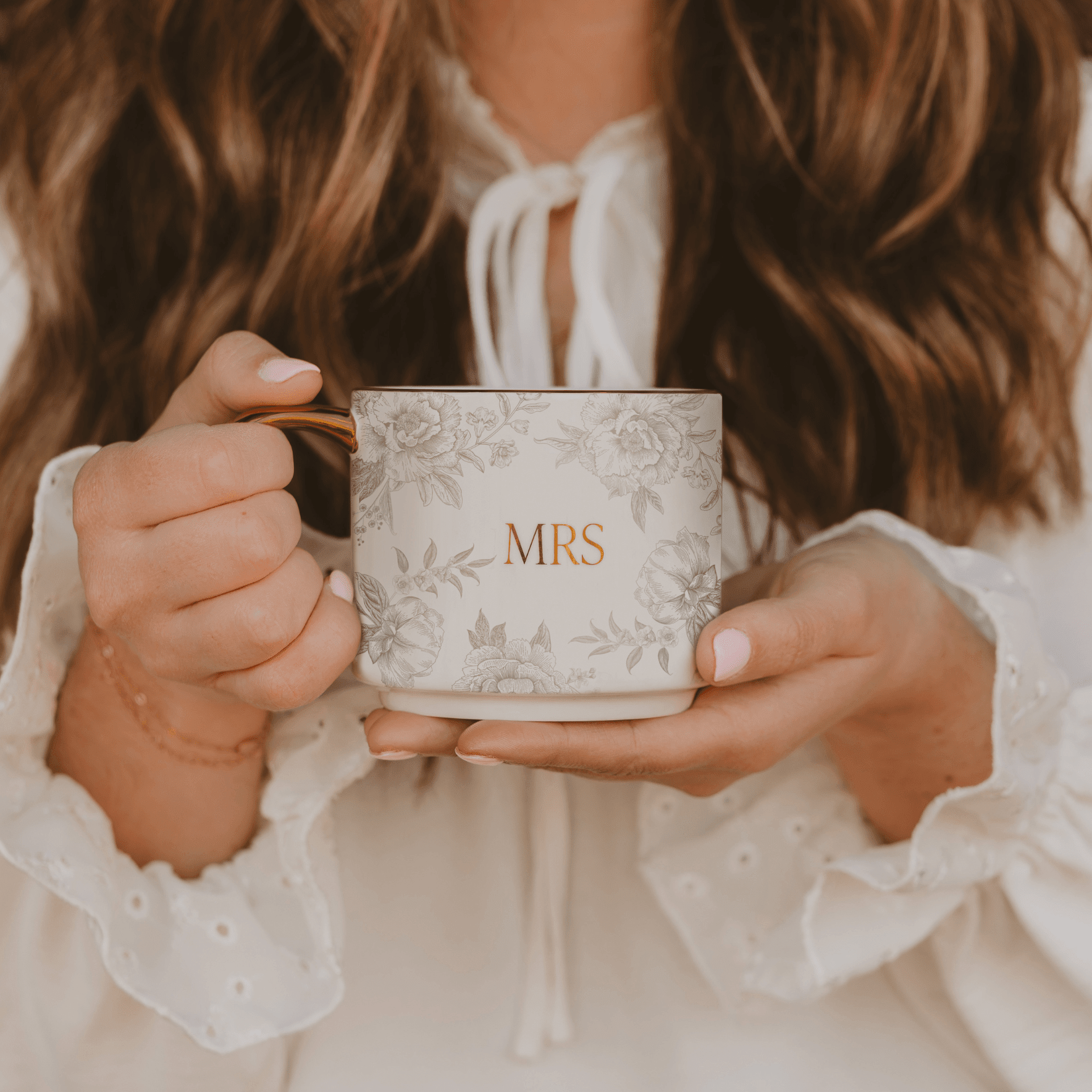 White ceramic mug with delicate floral pattern and gold "MRS" text, held by woman in white blouse