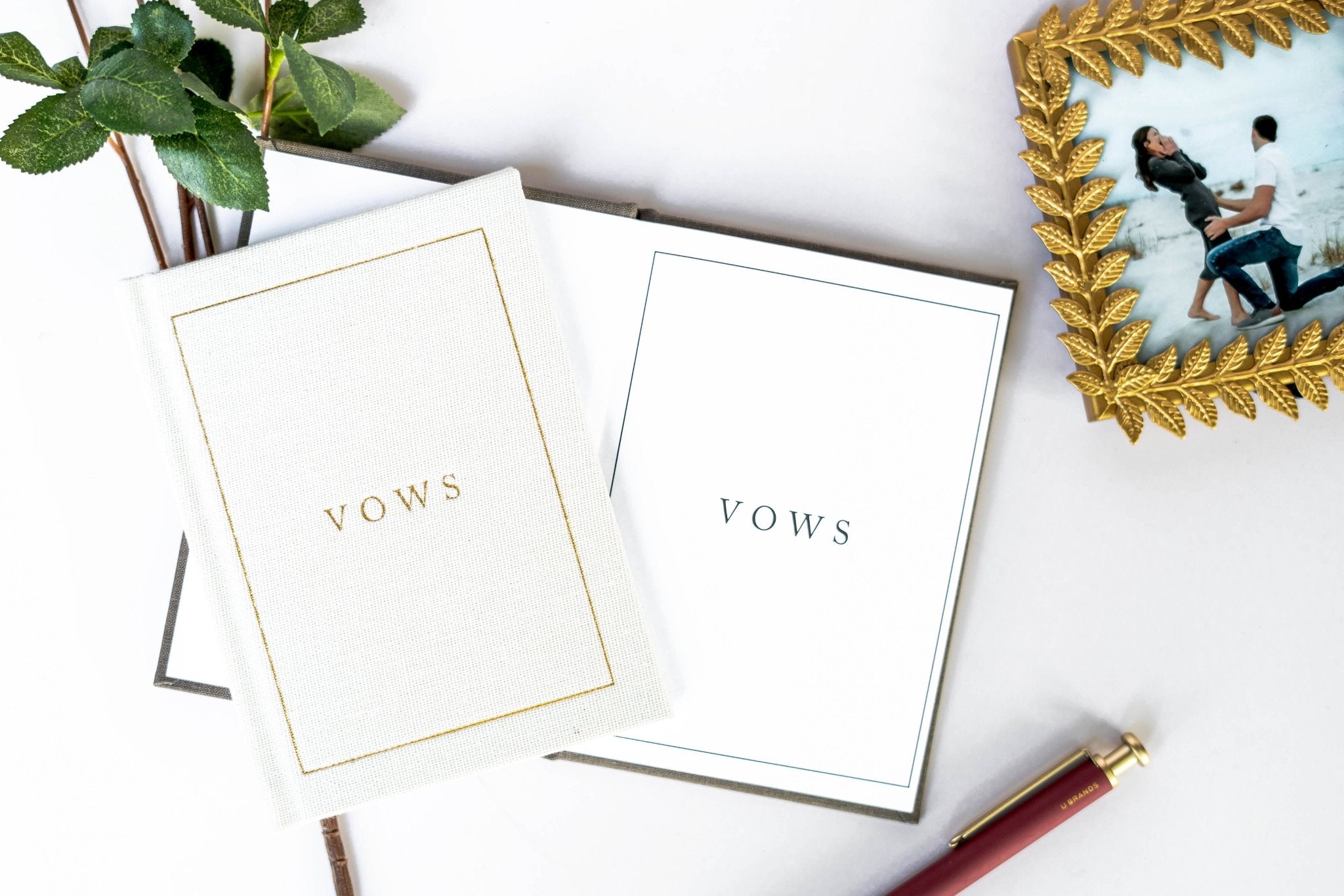 Two cream and white vow books with gold and gray borders, displaying "VOWS" text, arranged with a pen and greenery.