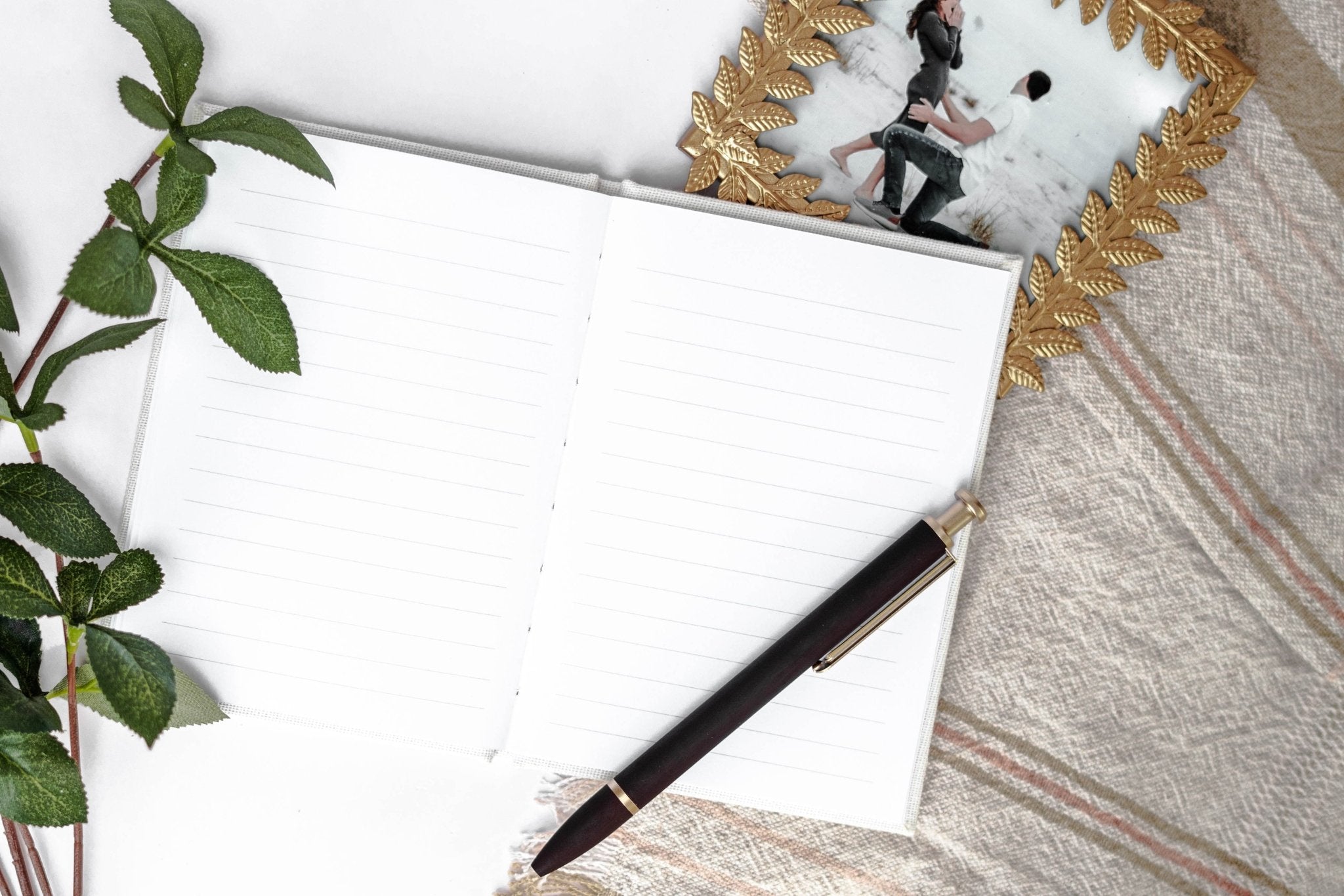 Blank white vow book with black pen, styled flat lay with green plant and striped fabric backdrop.