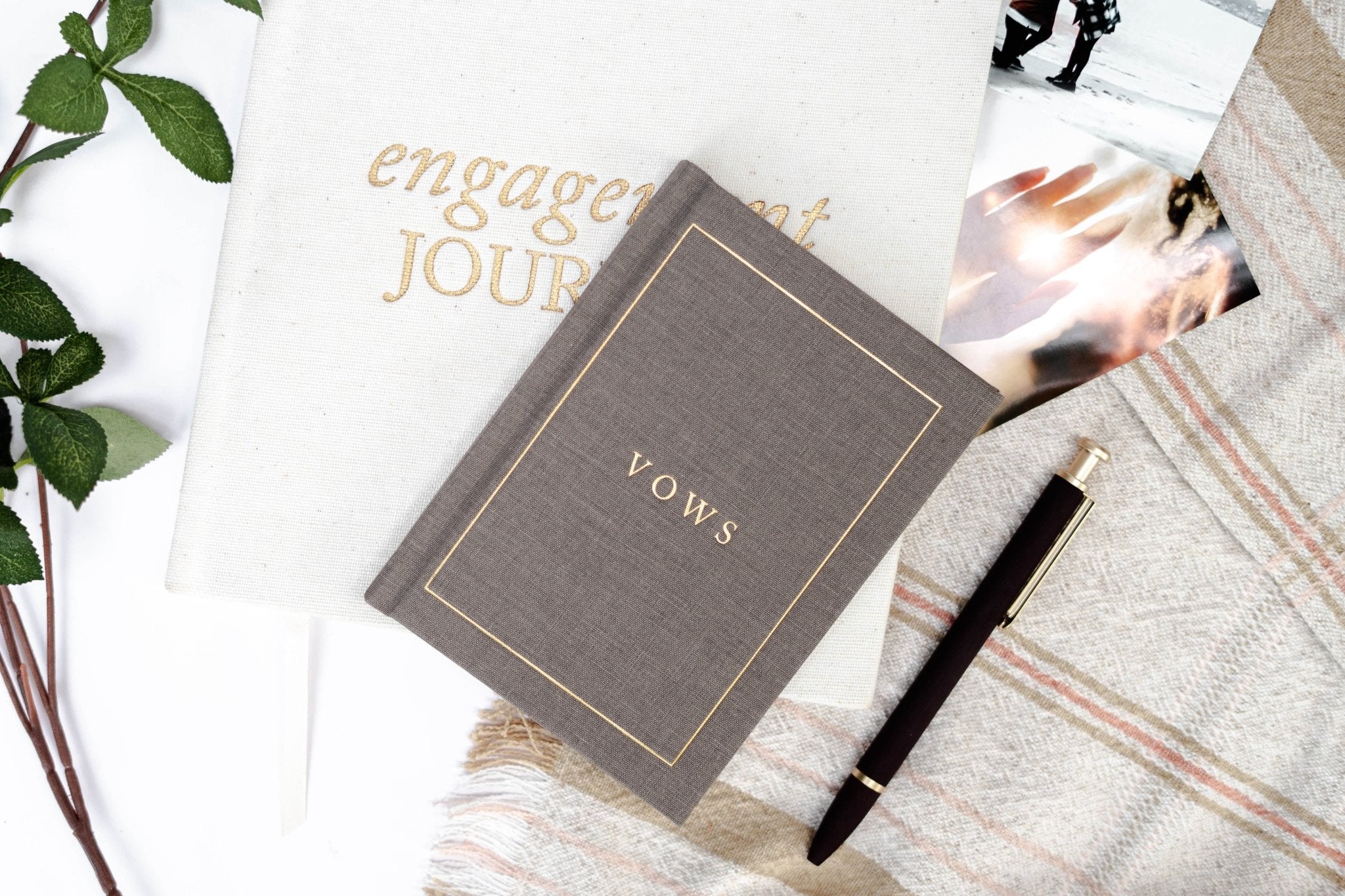 Gray linen vow book with gold "VOWS" text, styled flat lay with engagement journal, pen, and greenery.
