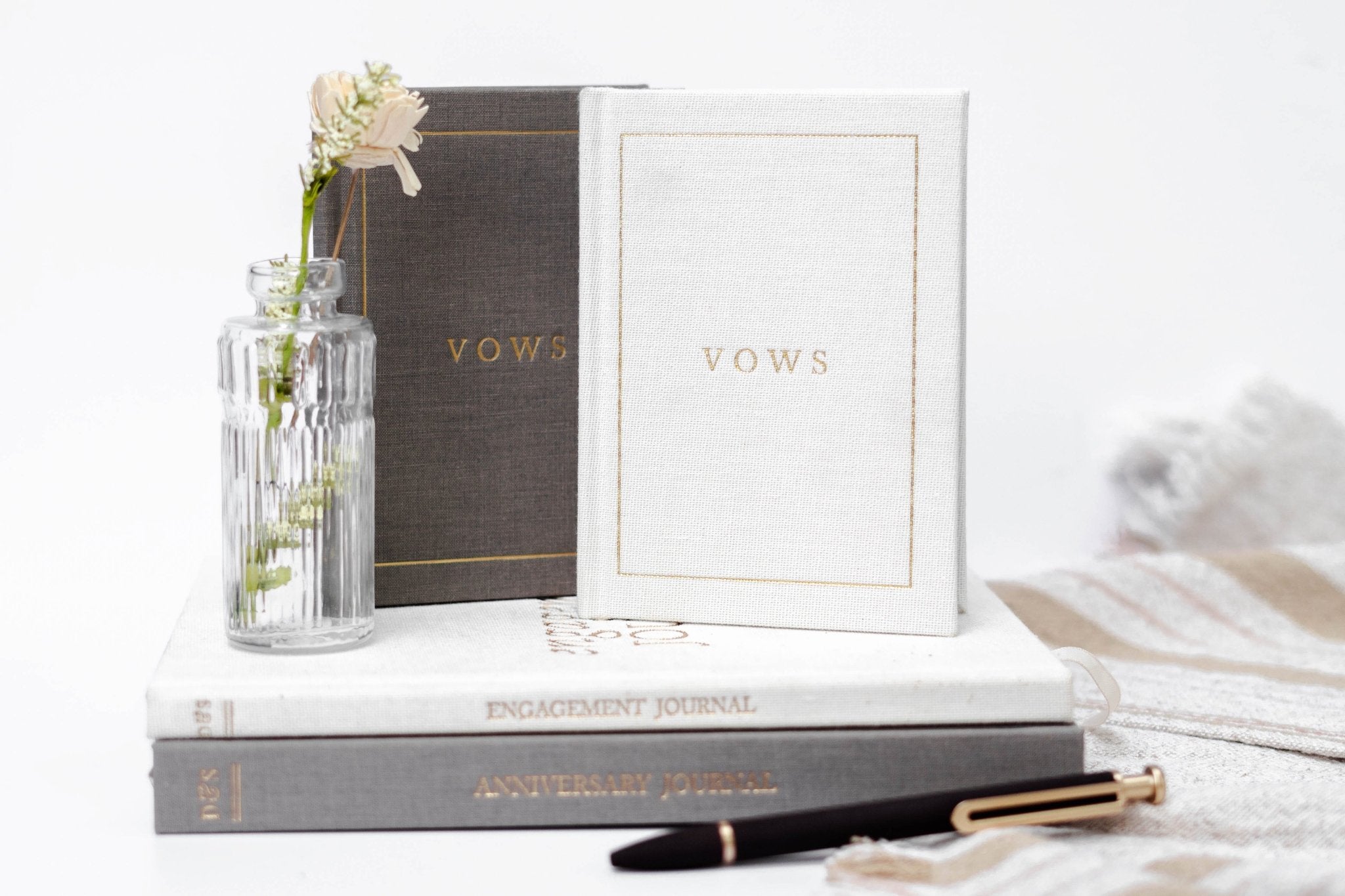 White and charcoal vow books with gold lettering stacked on books with a pen and small vase of flowers.
