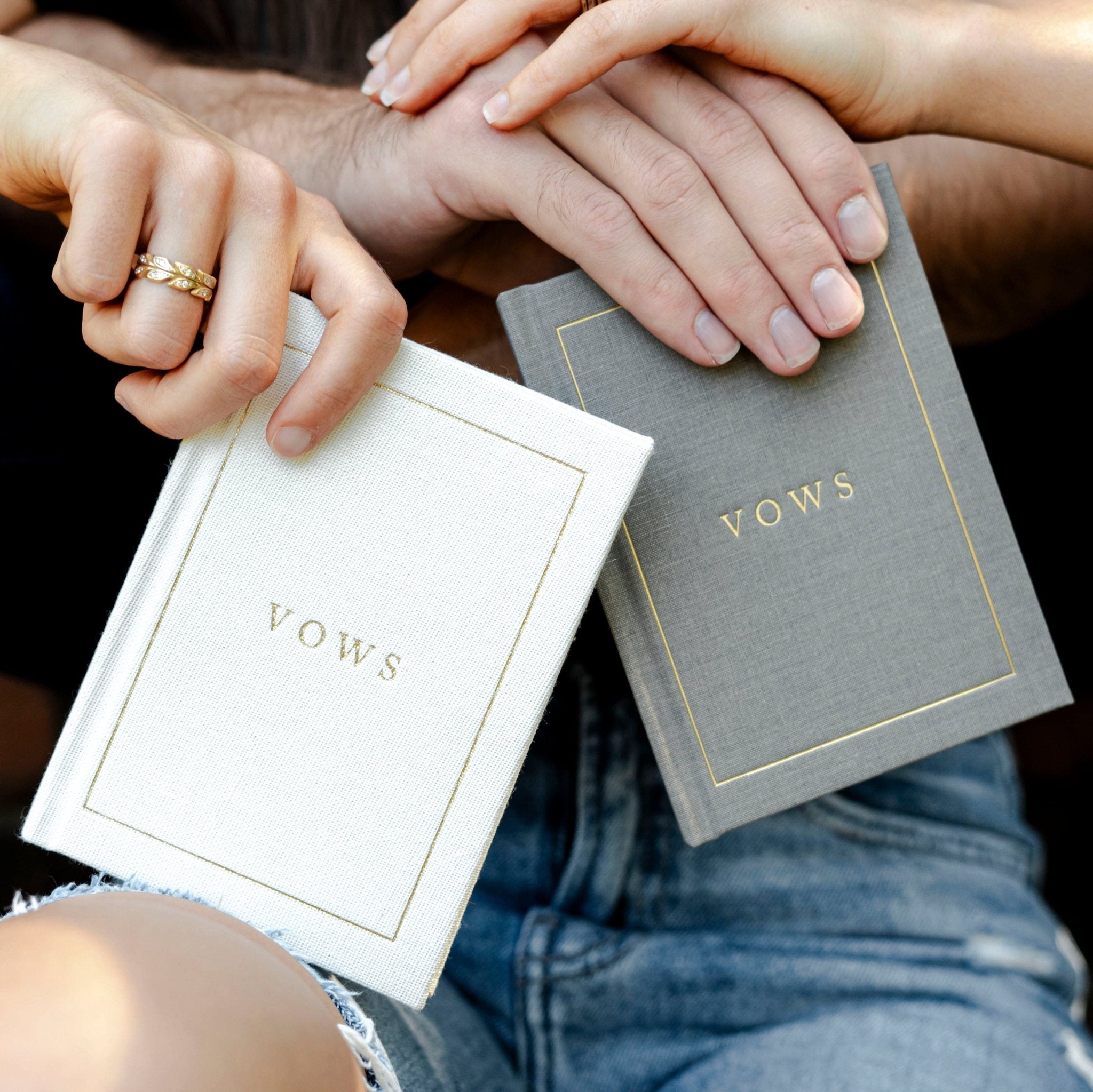 Couple holding white and gray vow books with gold "VOWS" text, displaying personalized wedding journal covers.