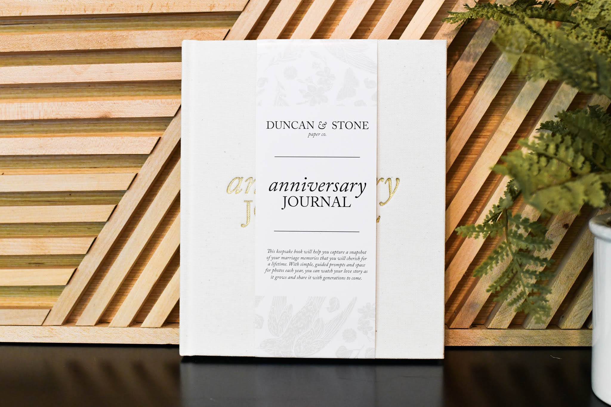 White anniversary journal with "Duncan & Stone" text and gold foil accents on wooden striped background