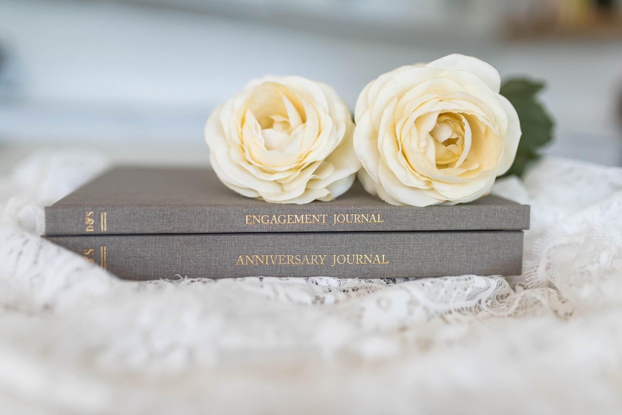 Cream roses resting on stacked gray anniversary and engagement journals with embossed gold lettering on white lace.