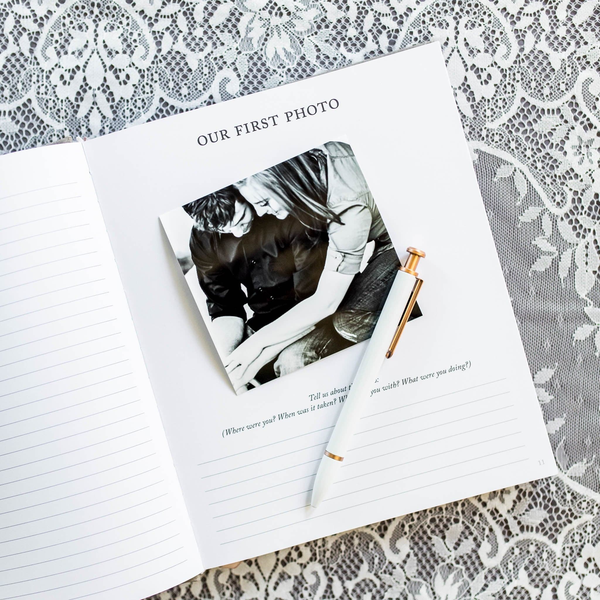 White anniversary journal with "Our First Photo" window cutout displaying couple's photo, lined pages, and gold pen on lace background.