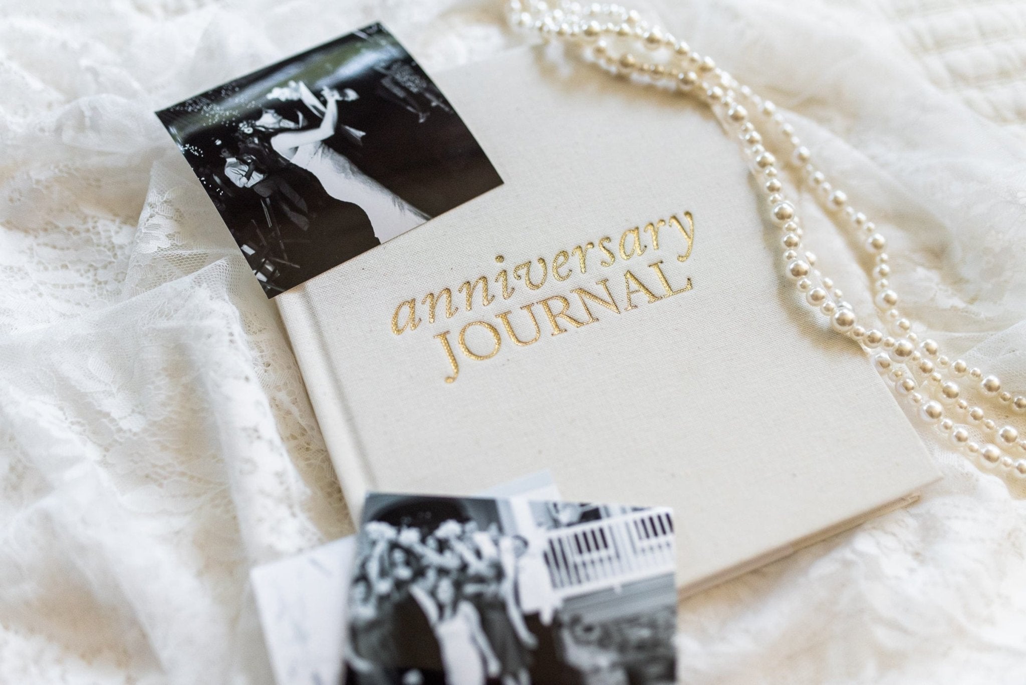 Cream-colored anniversary journal with gold foil text, displayed with black and white wedding photos and pearl beads on white lace fabric.