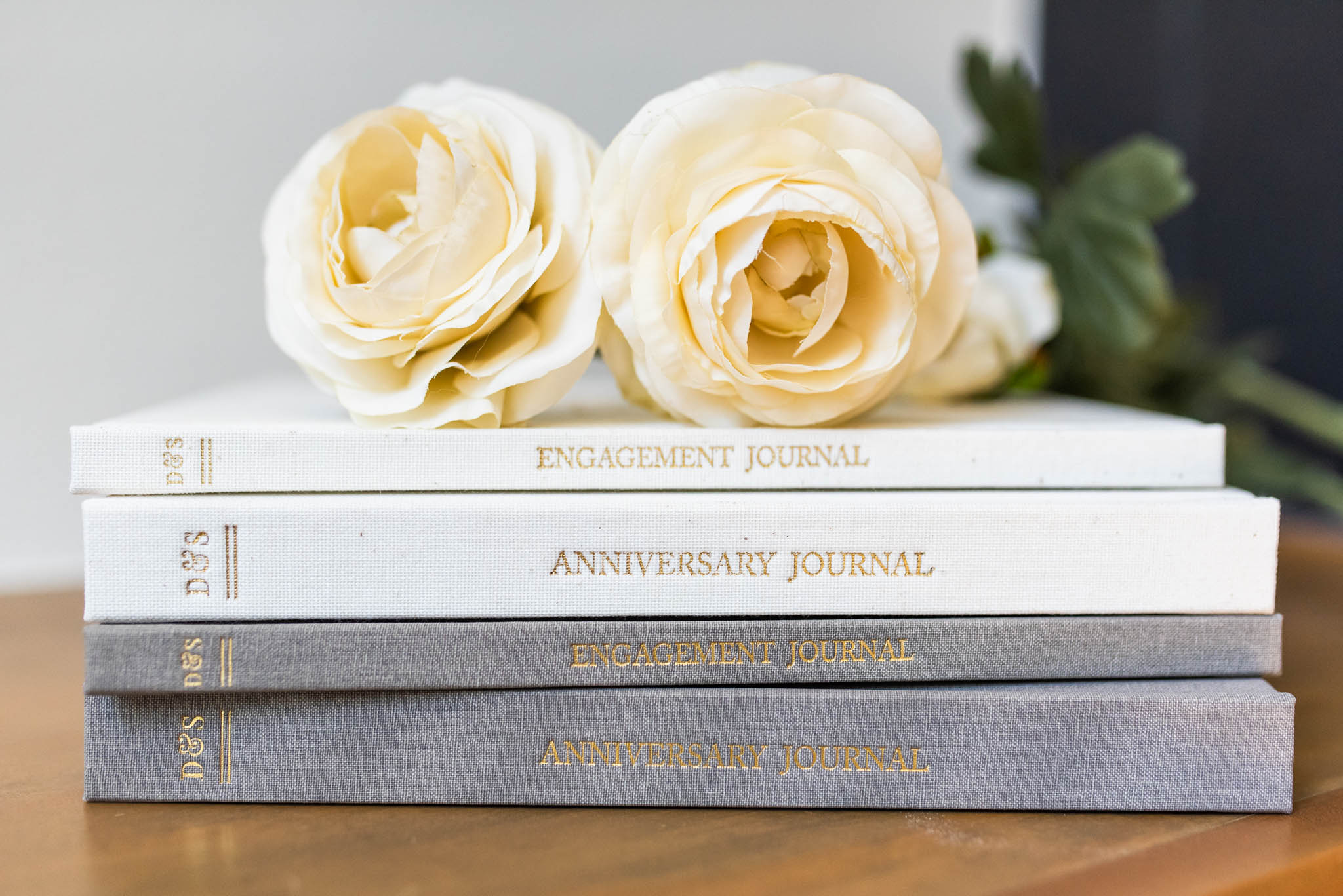 Stack of three anniversary and engagement journal books with gold foil text, topped with cream-colored roses