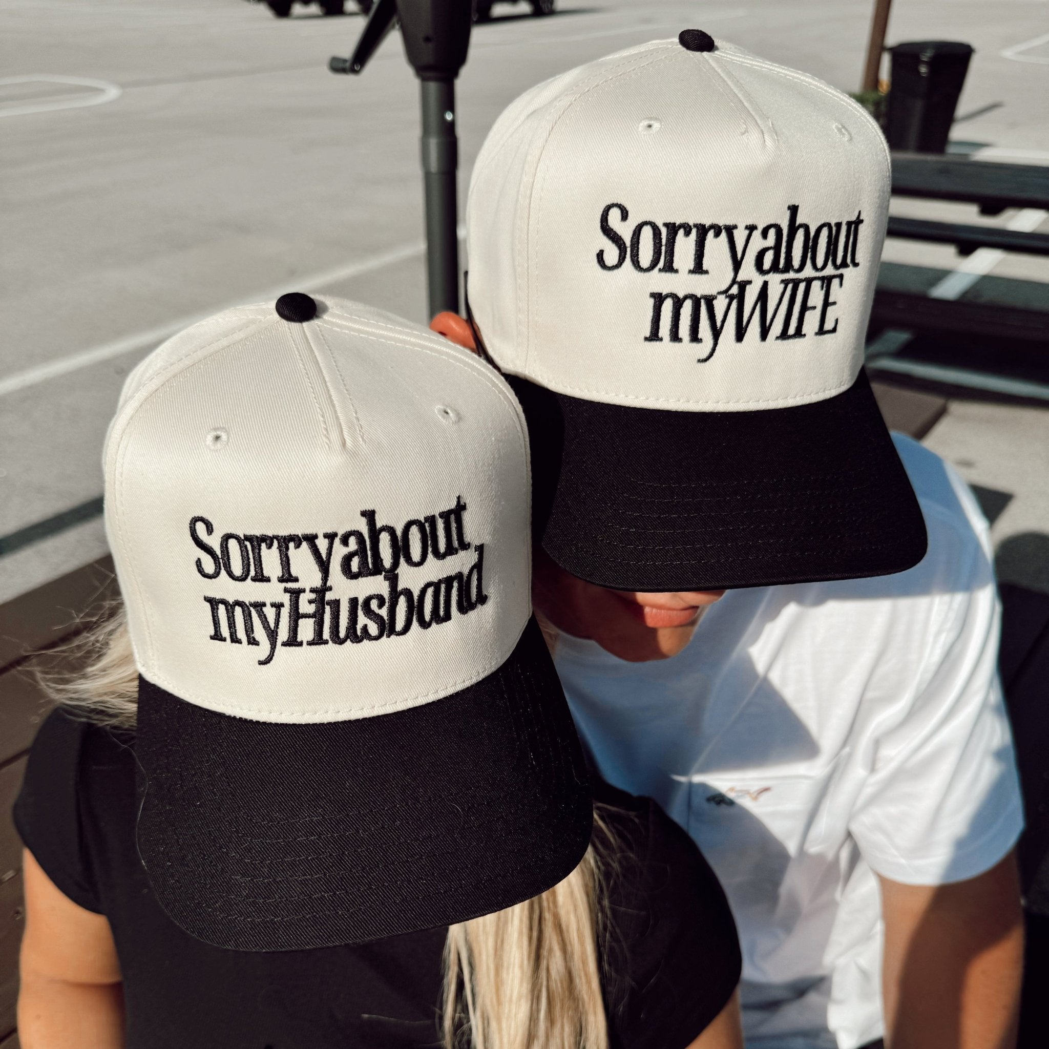 Pair of white and black trucker hats with "Sorry about my Husband" and "Sorry about my Wife" text on the front panels.