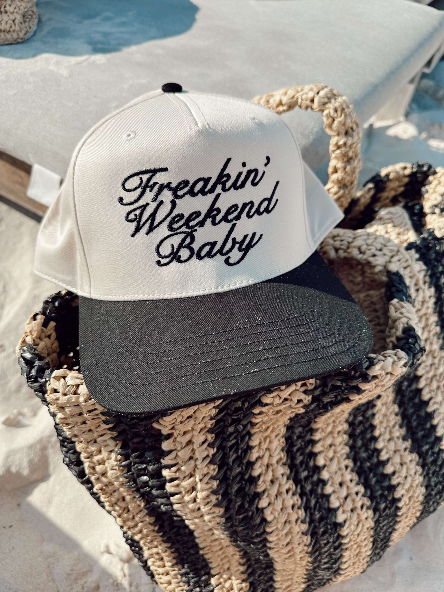 White and charcoal trucker hat with "Freakin' Weekend Baby" script text, displayed on striped beach tote by poolside.
