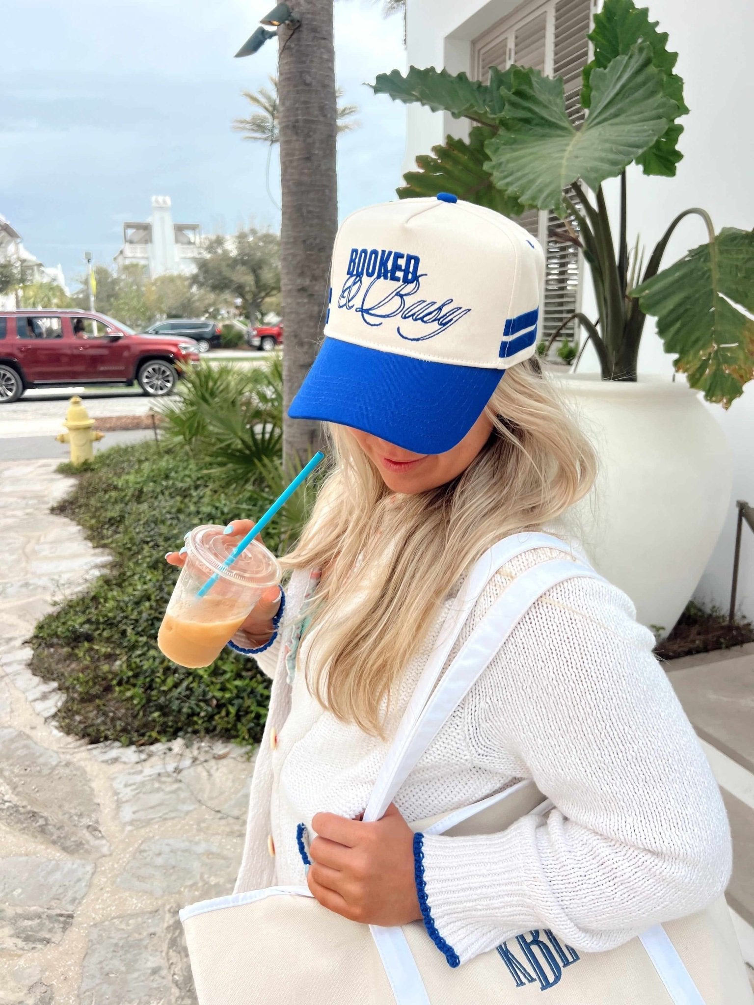 Cream and royal blue vintage trucker hat with "Booked and Busy" text, worn by woman holding beverage outdoors