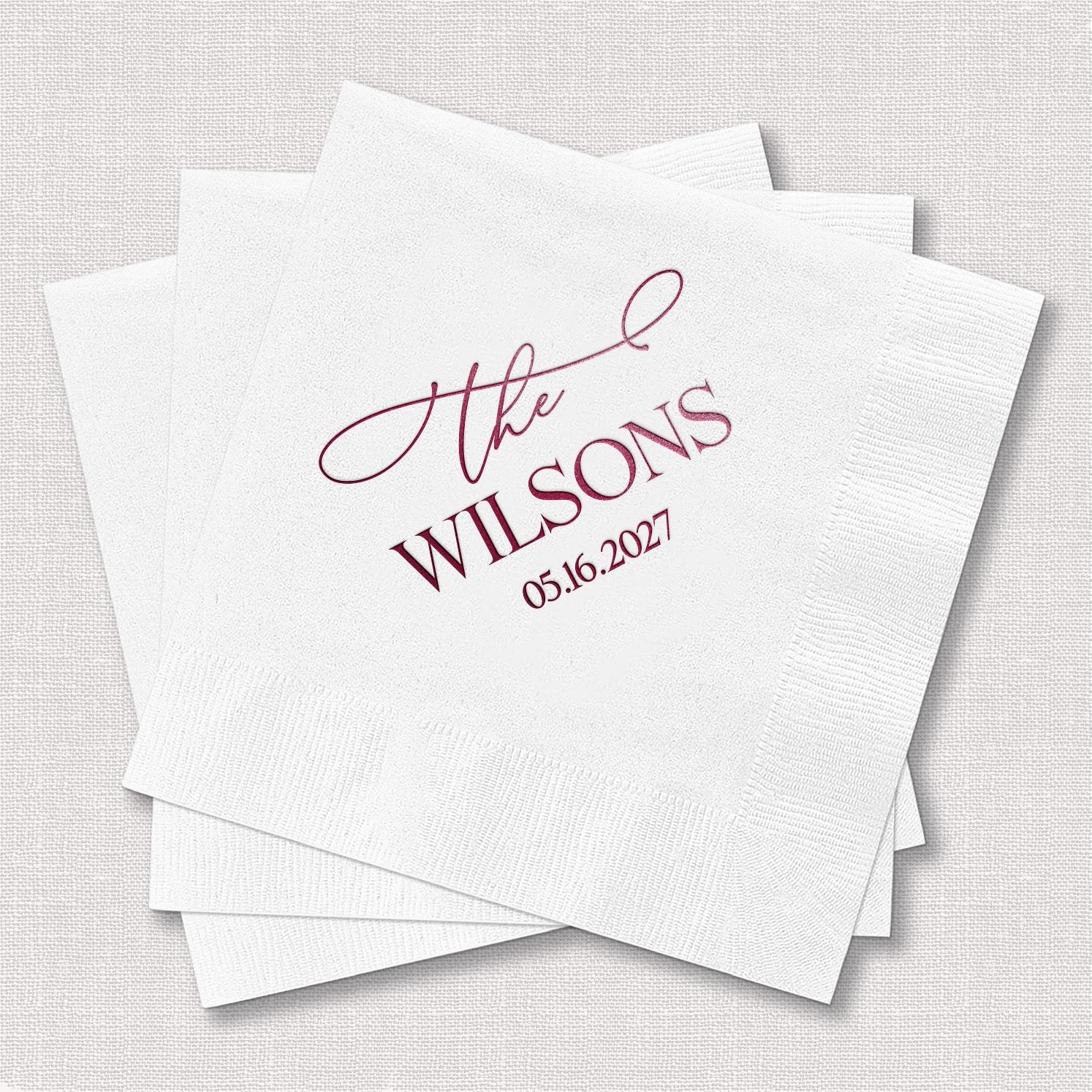 Stack of personalized white cocktail napkins with Elegant Last Name design featuring bold serif typography and delicate script in berry wine tones